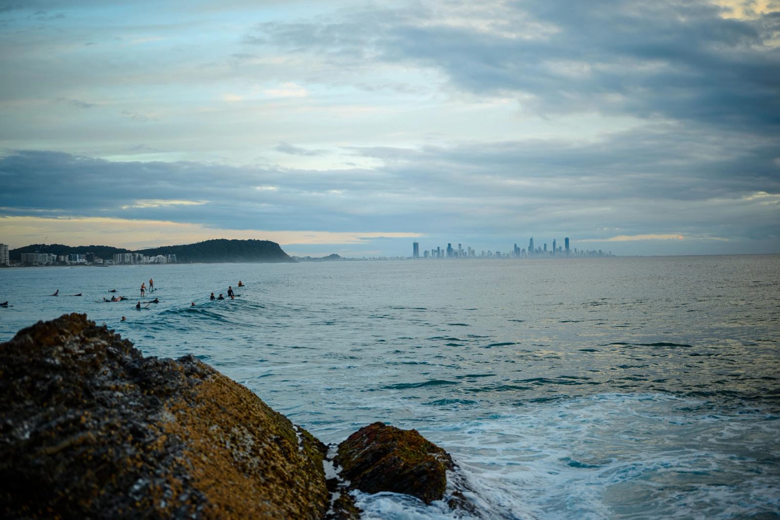 Surfers catching waves with a view of Gold Coast skyline from Currumbin Beach during sunset