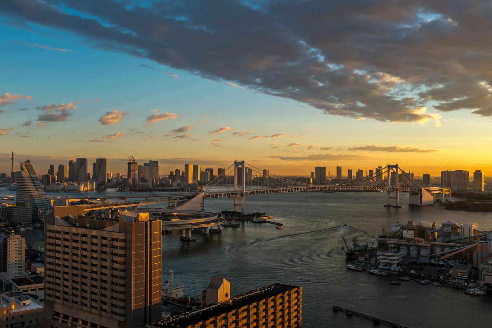 A suspension bridge crossing a harbour at sunset with a city skyline behind