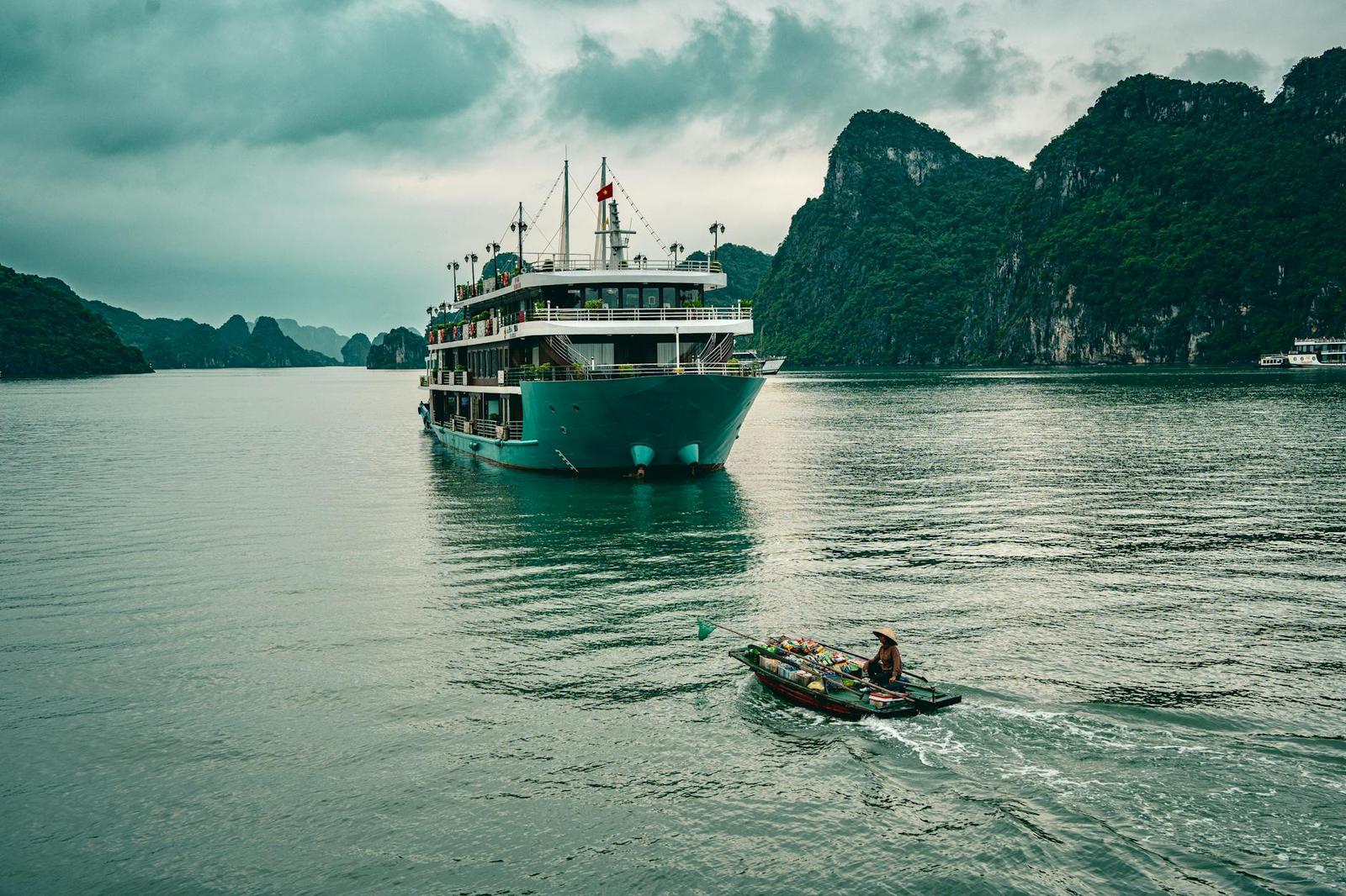 A small rowboat crossing in front of a large teal cruise boat on a misty bay