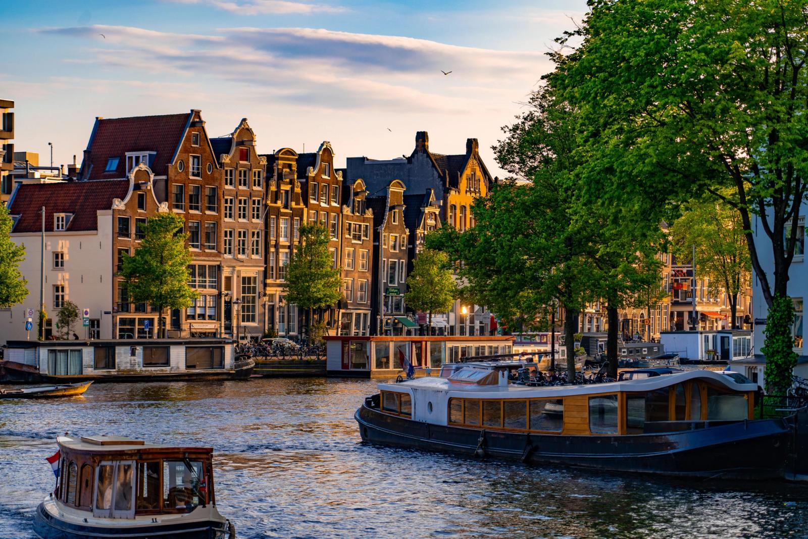 Scenic view of Amsterdam canal houses and boats at sunset with vibrant green trees