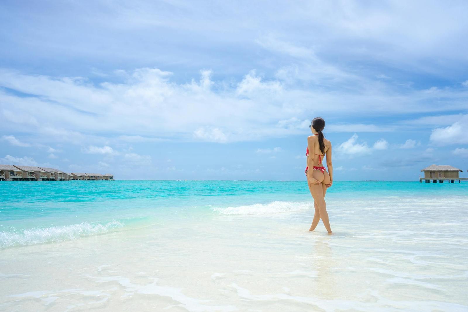 A woman in a bikini walking along a shallow lagoon beach near overwater villas