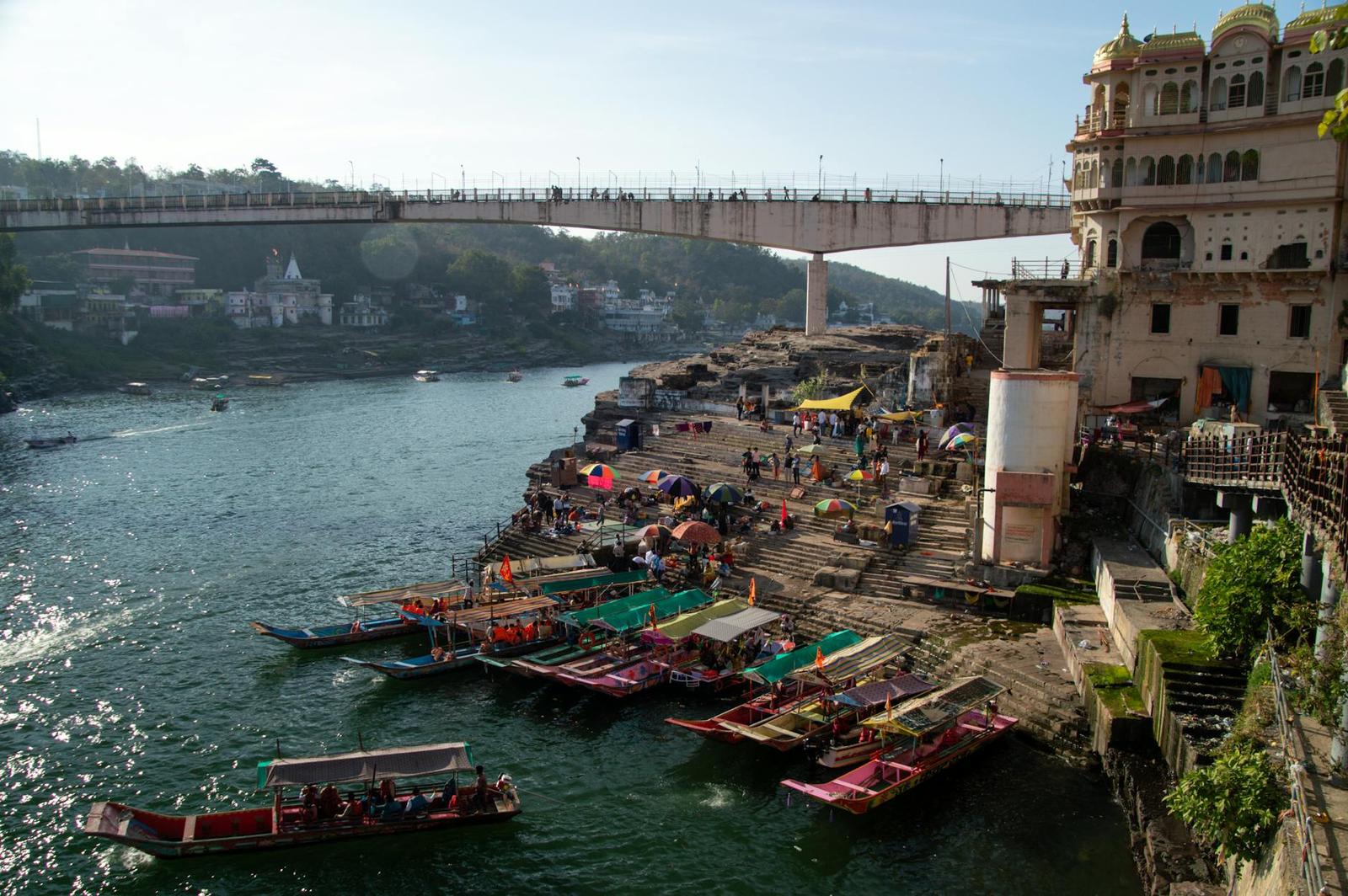 Riverside ghat with moored boats beneath a pedestrian bridge and temple