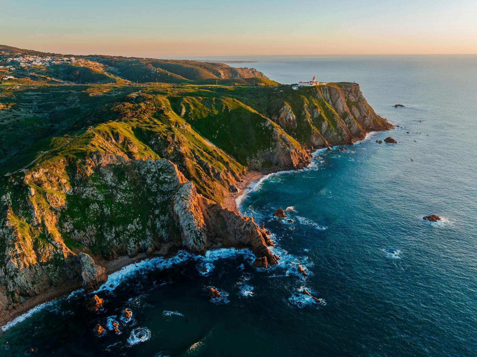 A lighthouse stands atop green cliffs overlooking the ocean at sunset