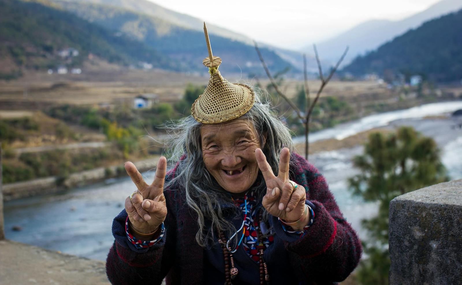 An elderly woman in traditional dress flashing peace signs beside a mountain river
