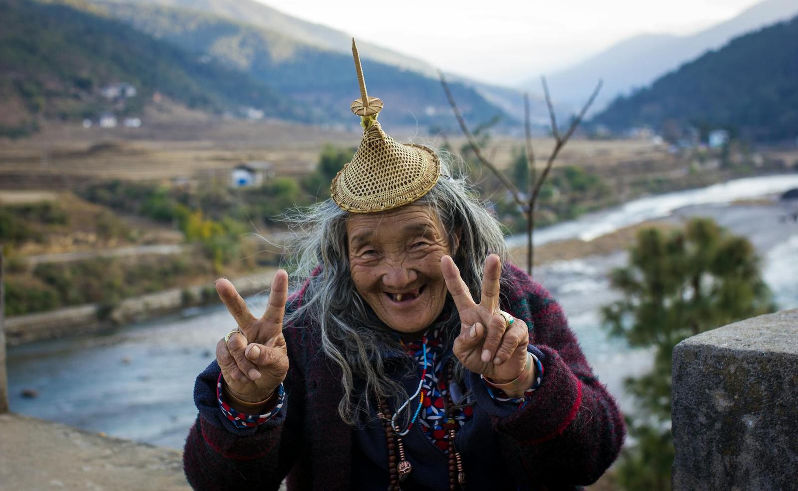 An elderly woman in traditional dress flashing peace signs beside a mountain river