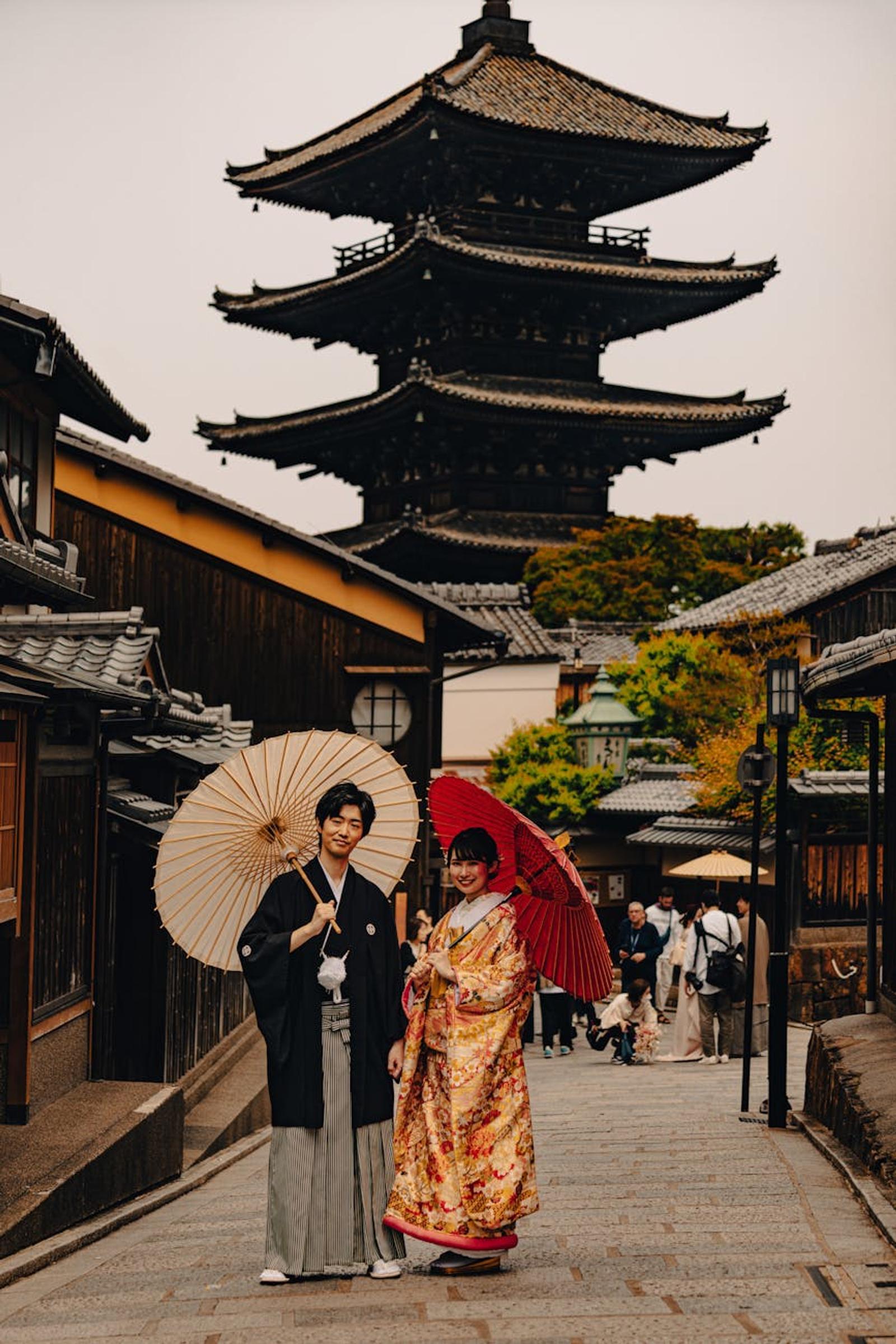 Couple in traditional kimono holding paper umbrellas on a lane before a wooden pagoda