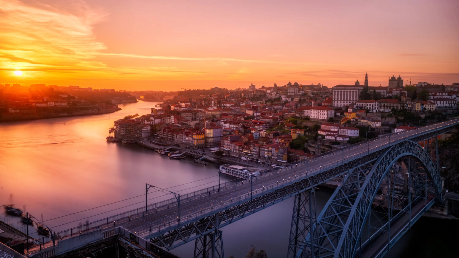 A riverside old town at sunset beneath a tall iron arch bridge