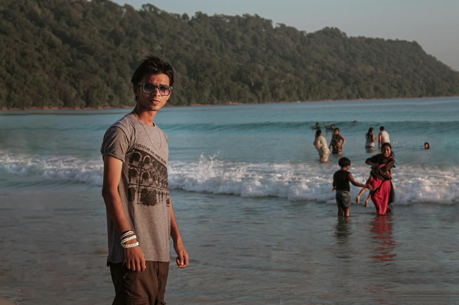 A young man in sunglasses on a beach with families wading in the surf behind him