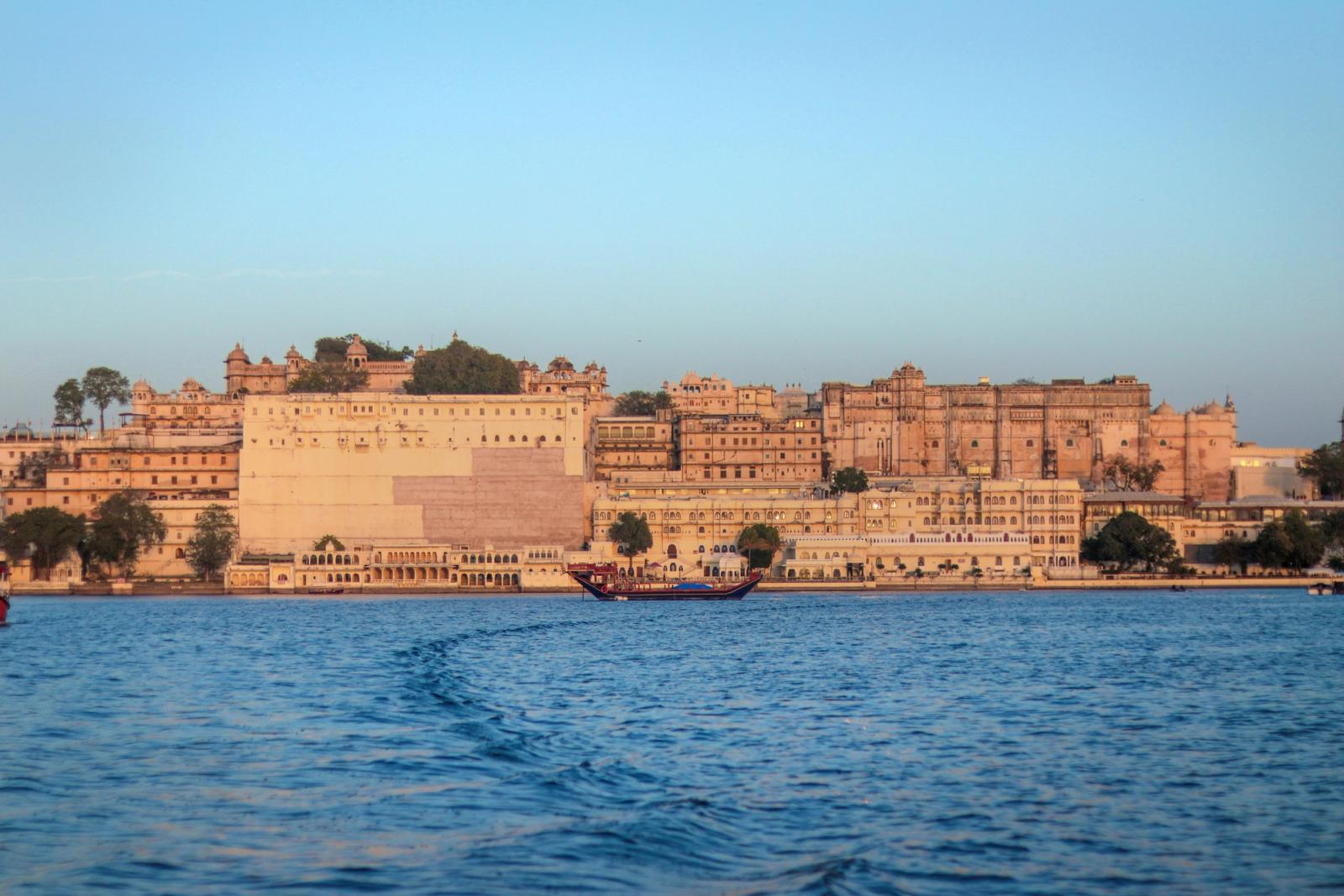 A sandstone lakefront palace complex with a boat trailing a wake at dusk