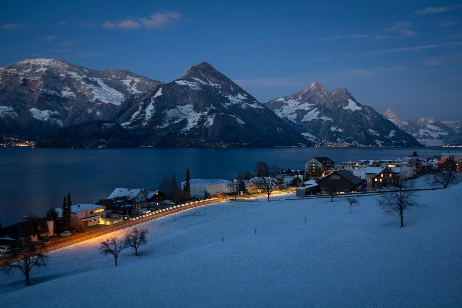 Lakeside village lit up at dusk beneath snow-covered peaks