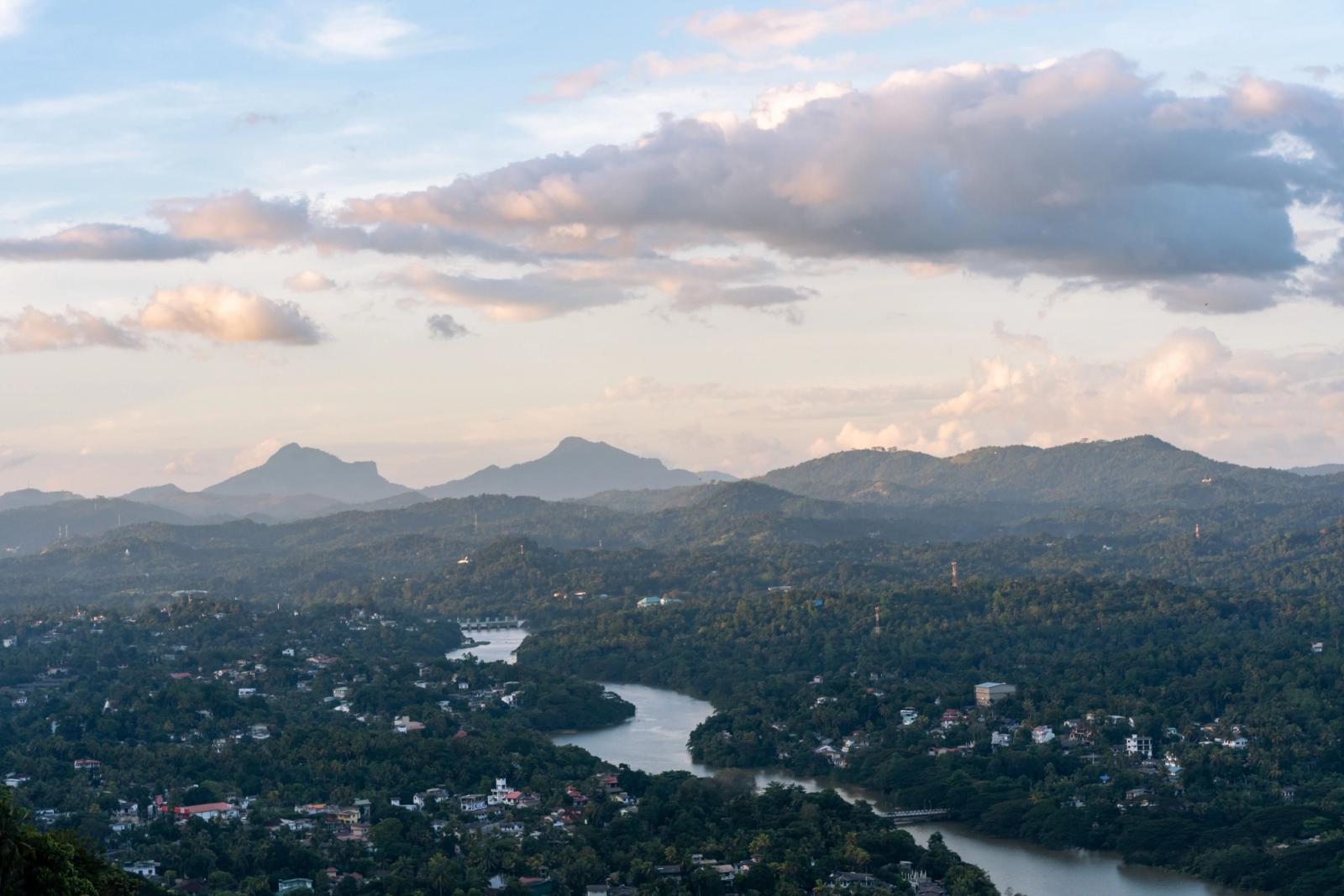 Aerial view of lush green Sri Lankan hills with a calm river and distant mountains under a cloudy sky