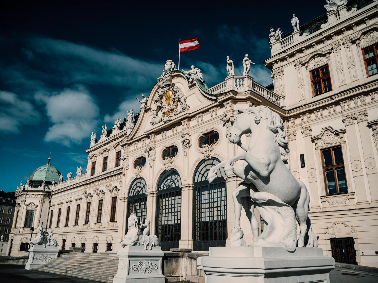 A baroque palace facade with a rearing horse statue and a flag against a dark sky