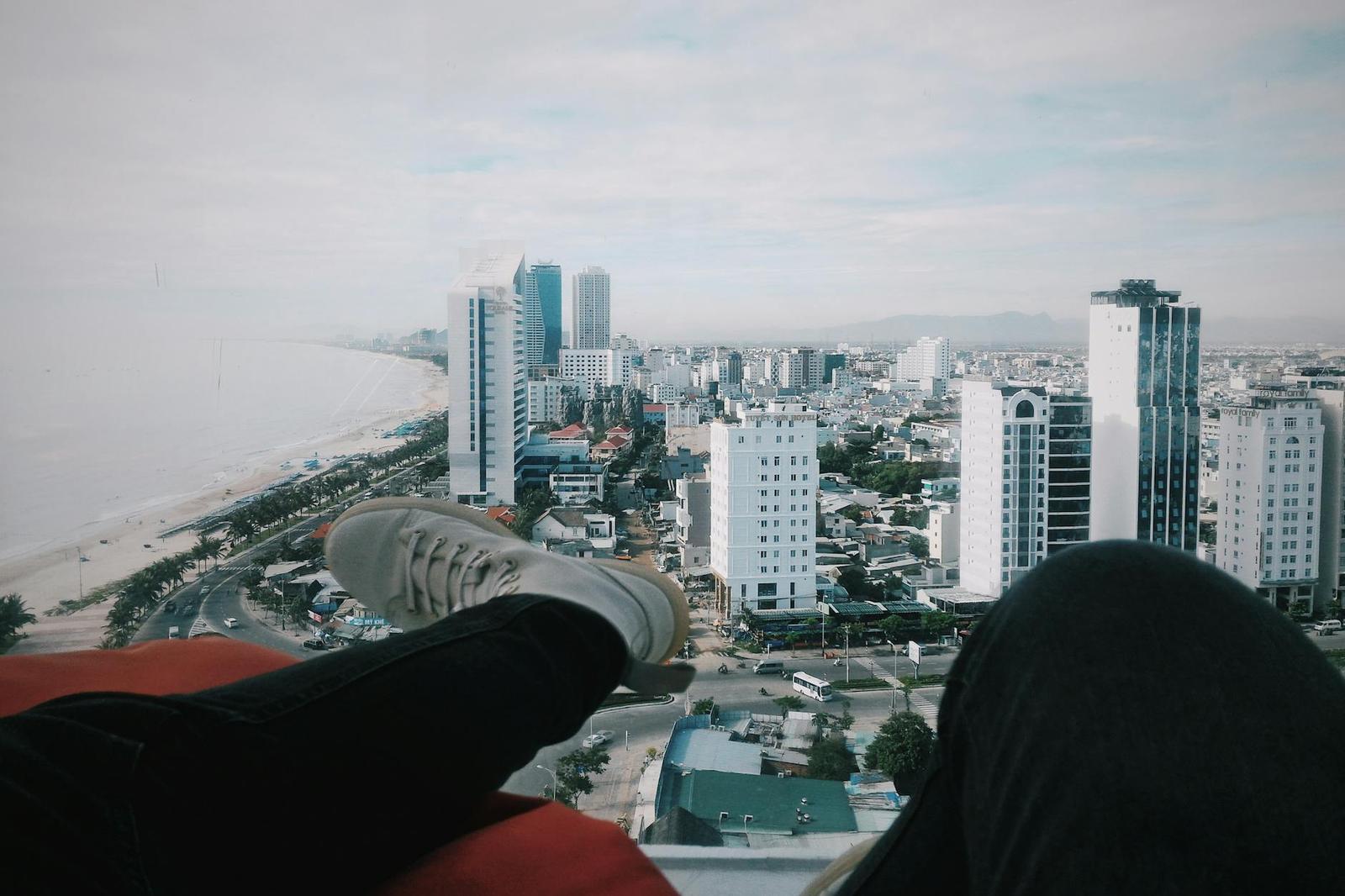 View over dangling feet toward a coastal city skyline and beach from a height