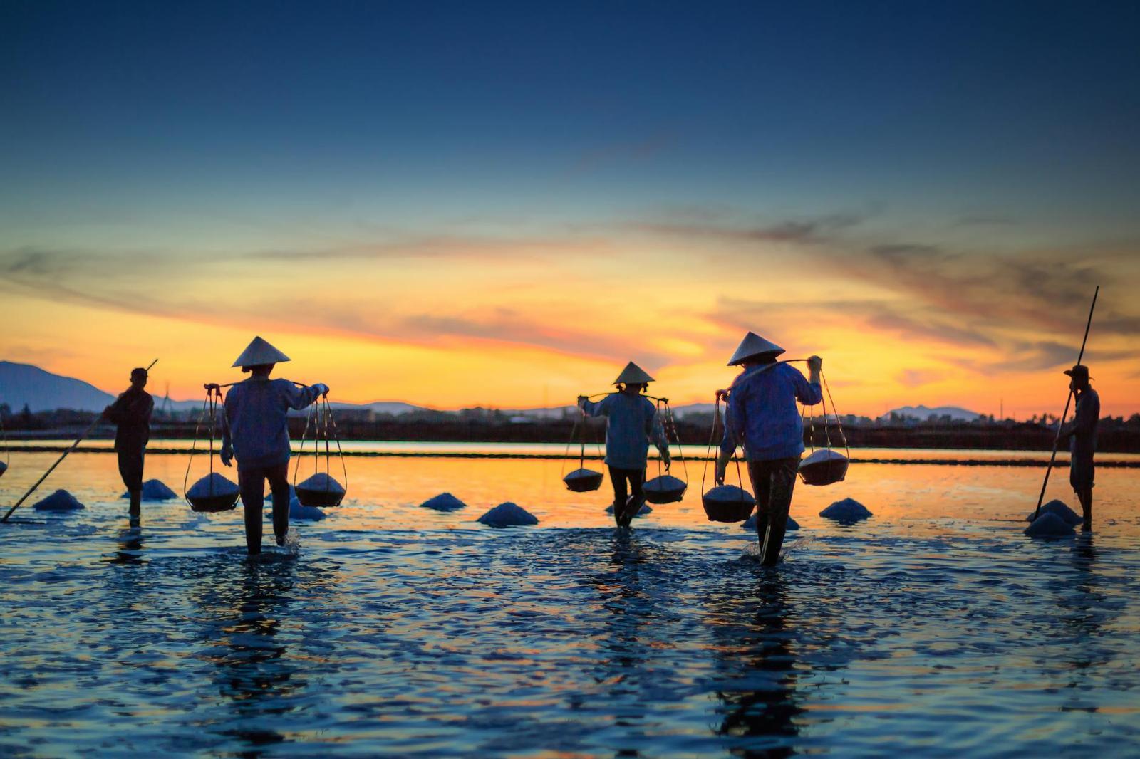 Silhouetted figures wading through shallow water carrying shoulder baskets at dusk