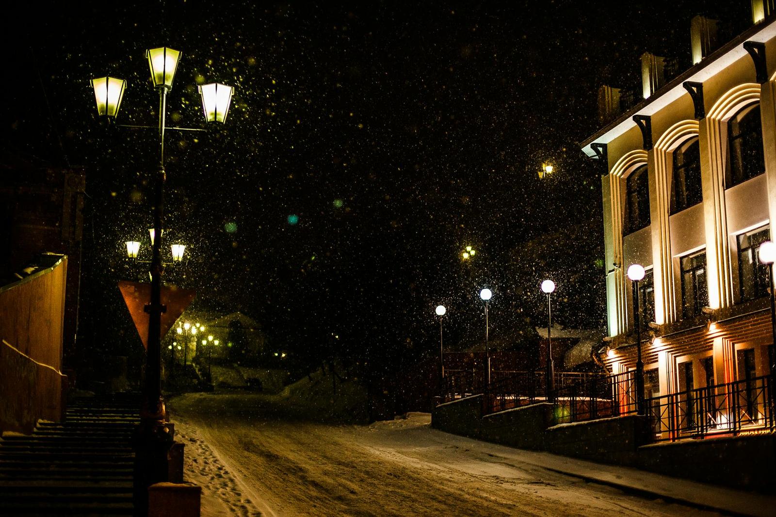 Snow falling on a lamplit street beside a lit building at night