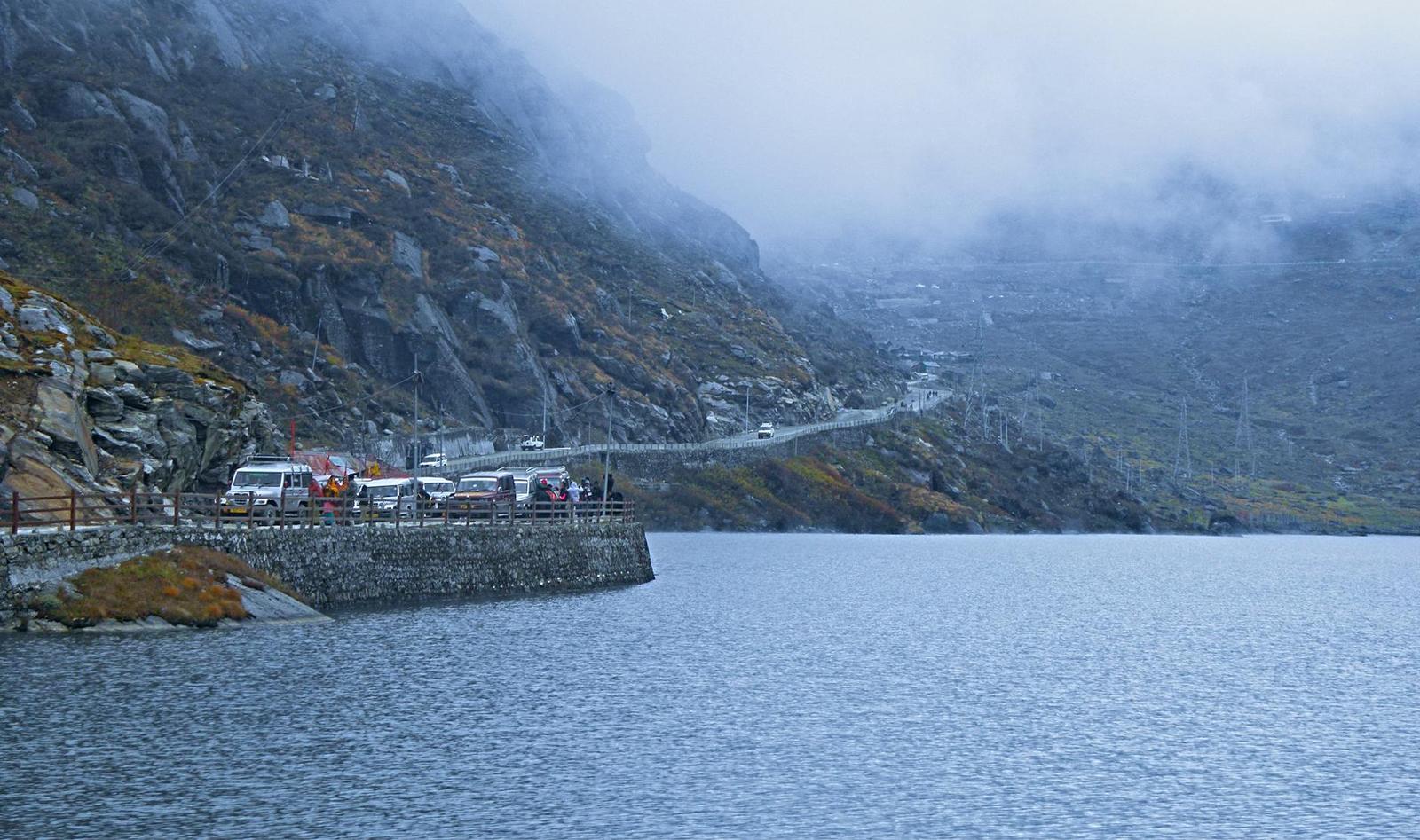 Parked vehicles beside a mountain lake with mist rolling over surrounding cliffs