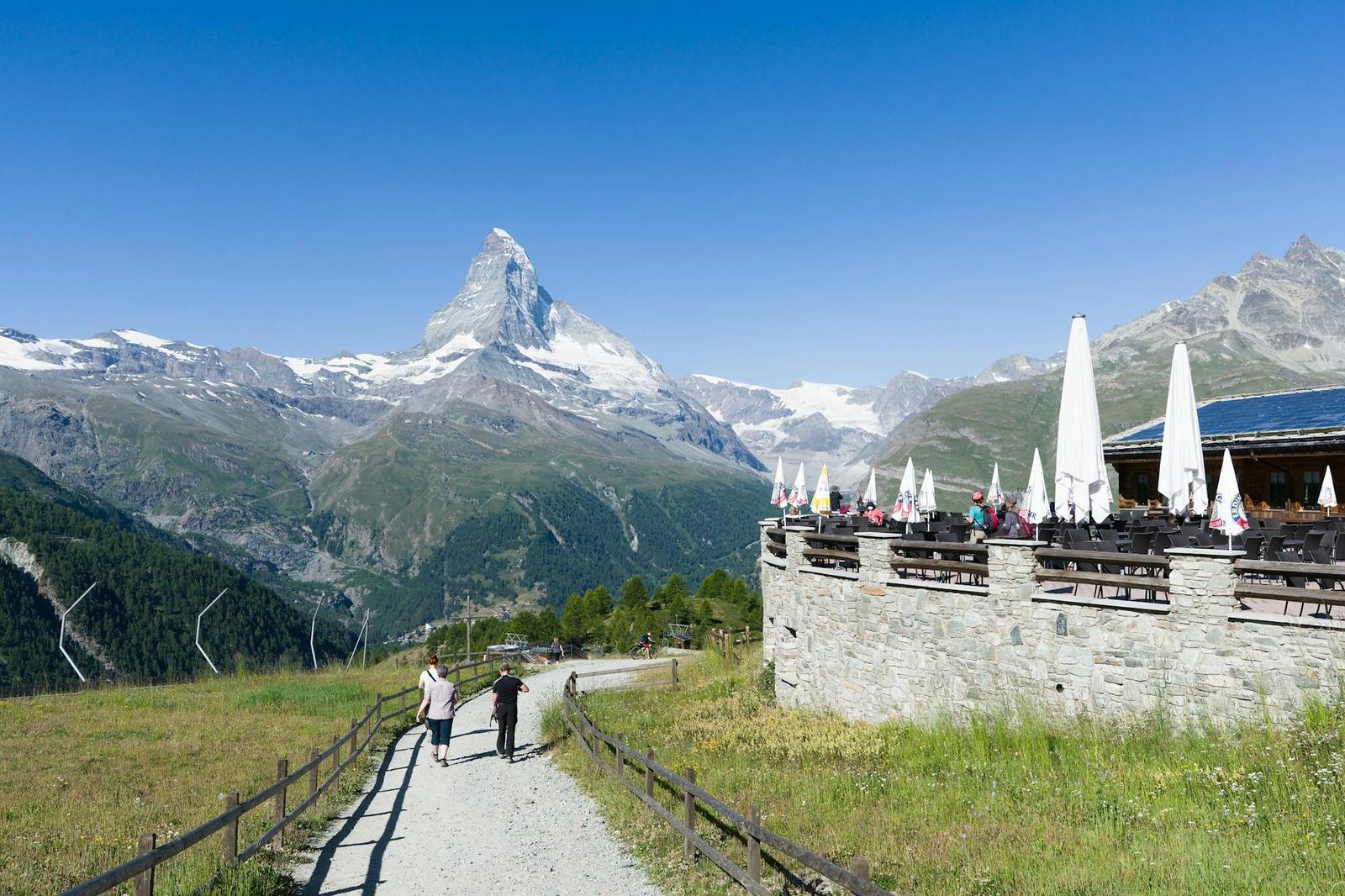 Hikers on a mountain path beside a stone terrace facing a pyramid peak