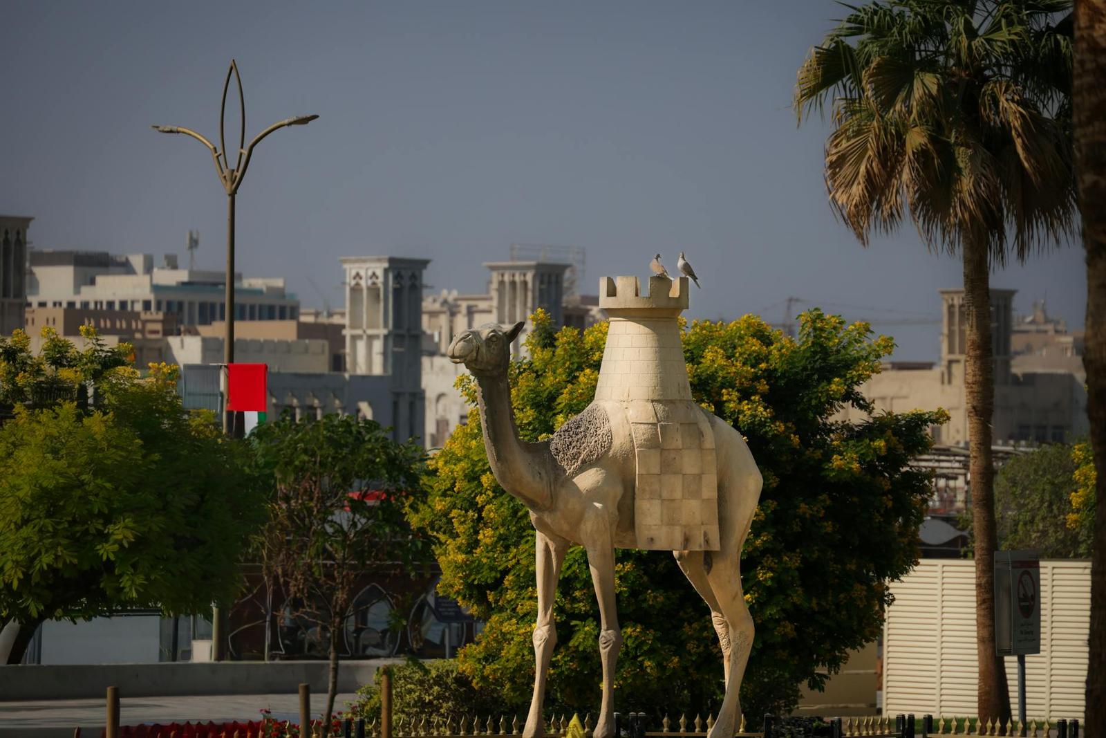 A camel statue surrounded by lush greenery and urban architecture under clear skies