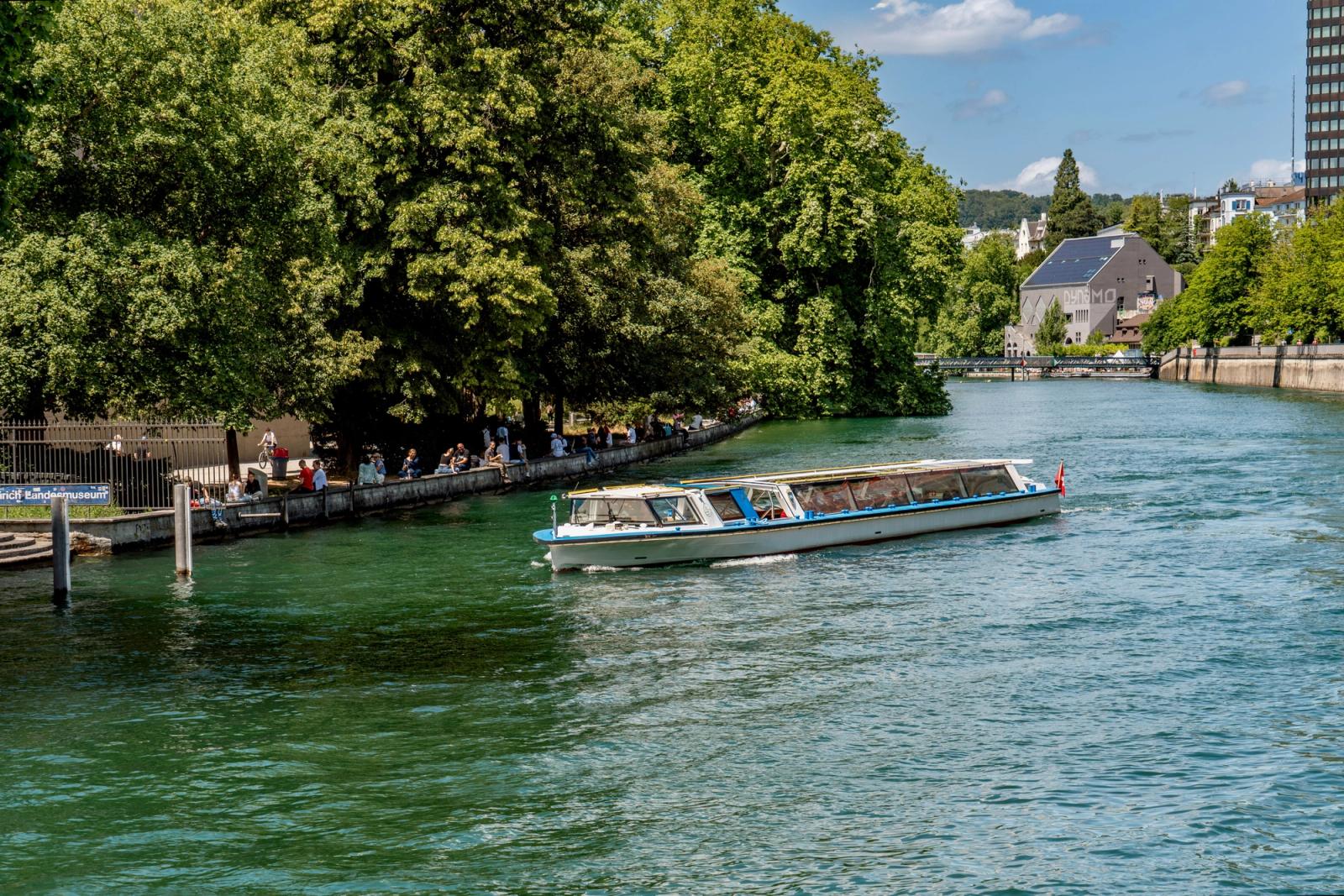 A tour boat cruises down a vibrant green river in Zurich, surrounded by lush greenery and urban architecture