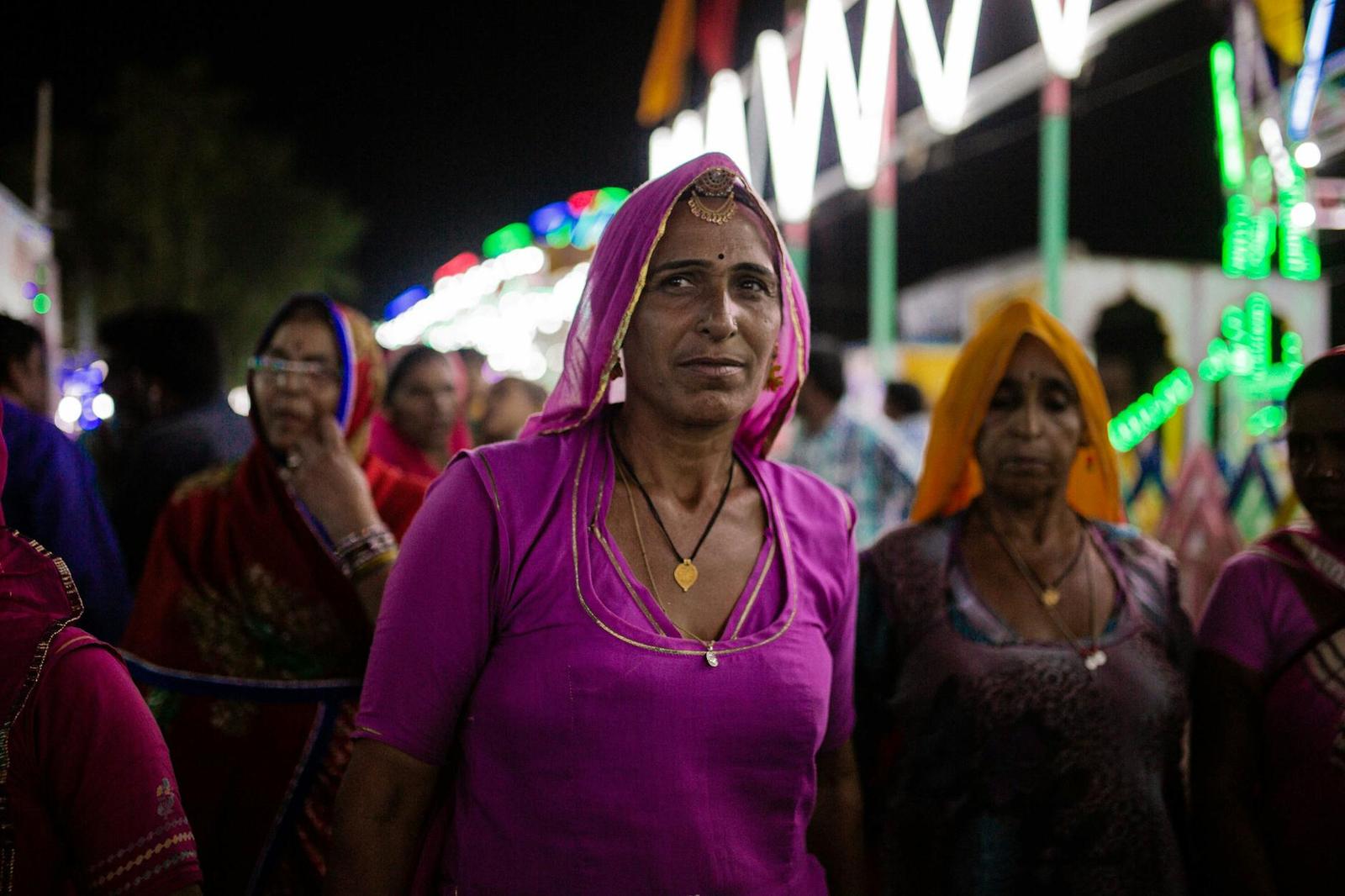 A close-up of a woman in a pink veil among a crowd at a lit fairground