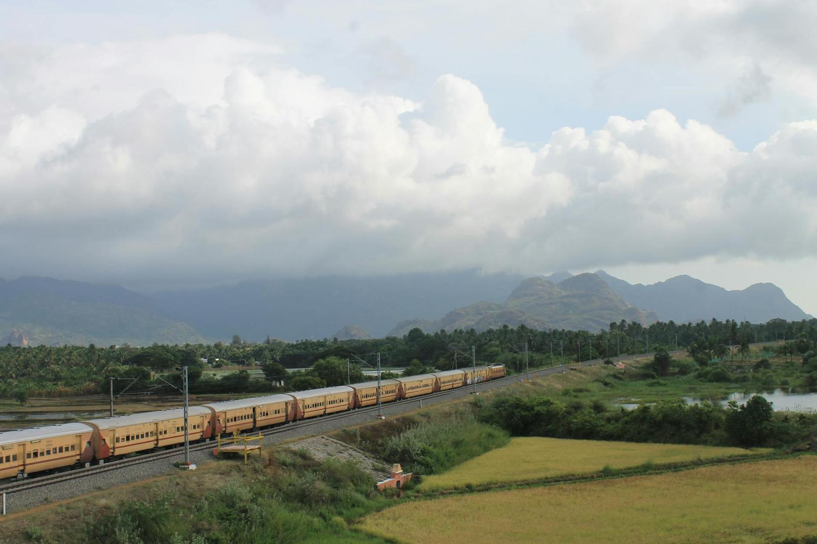 A long passenger train winding through green fields beneath cloud covered hills