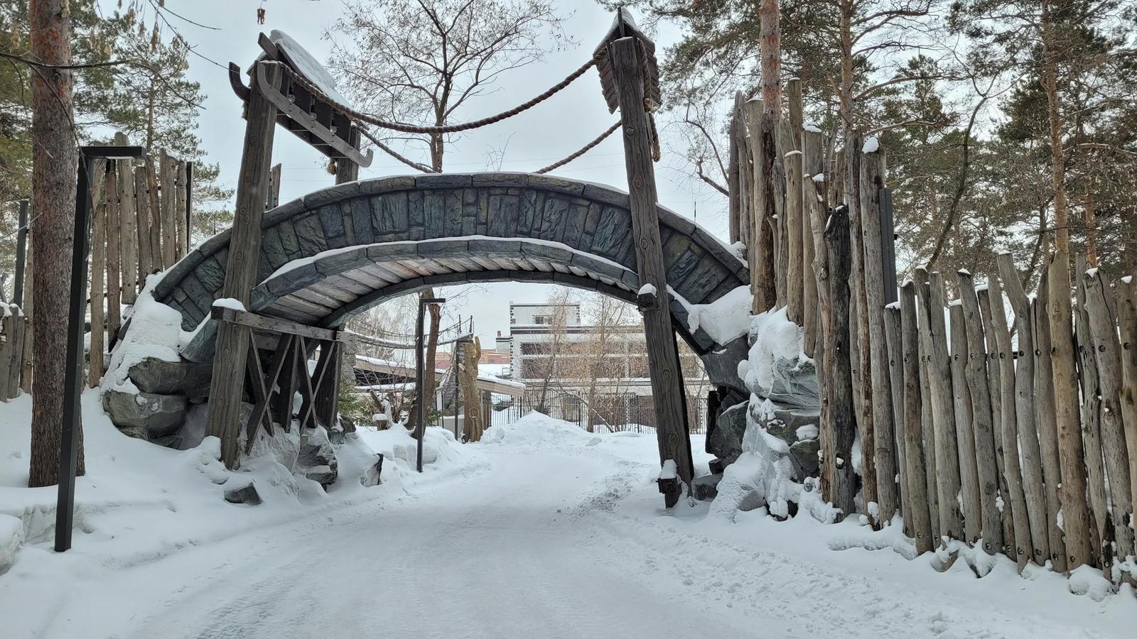 Snow-covered log archway over a path lined with wooden fencing