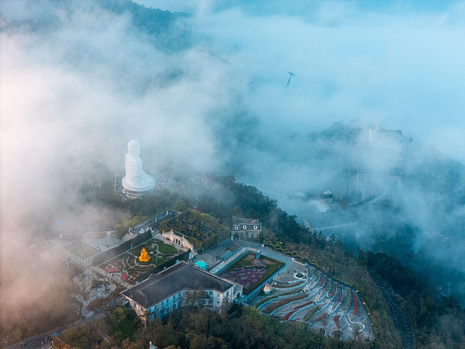 A tall white statue on a hilltop temple complex wreathed in mist