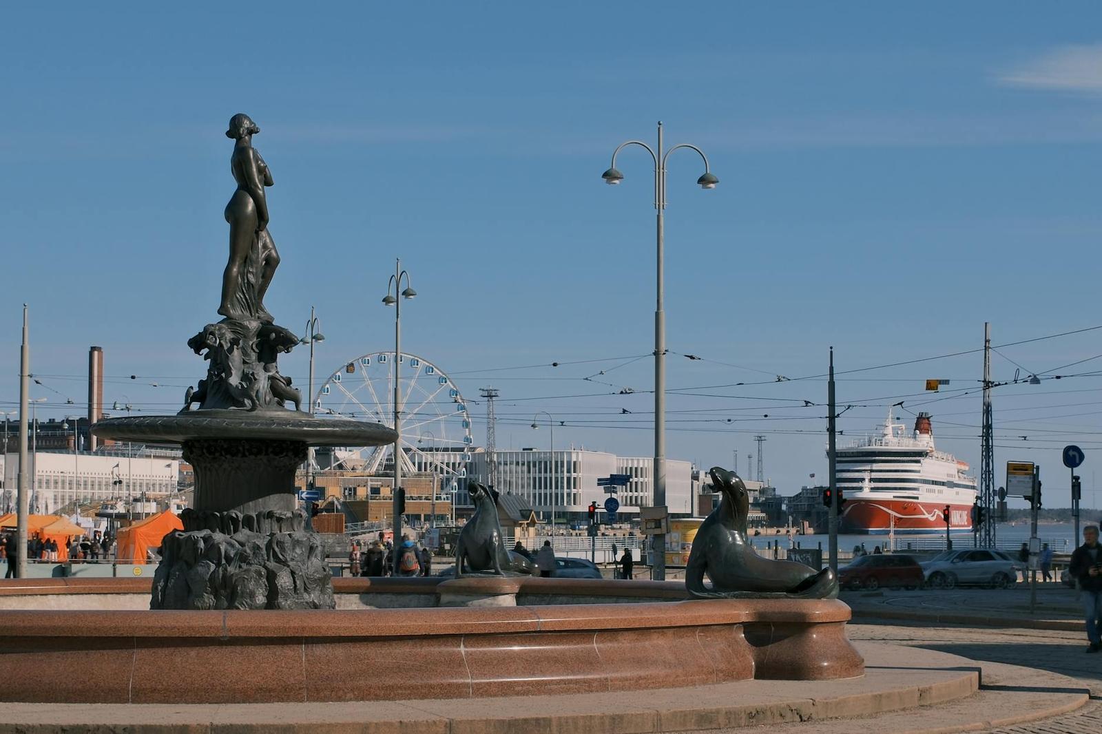 A bronze fountain statue overlooking a harbour with a ferris wheel and docked cruise ship