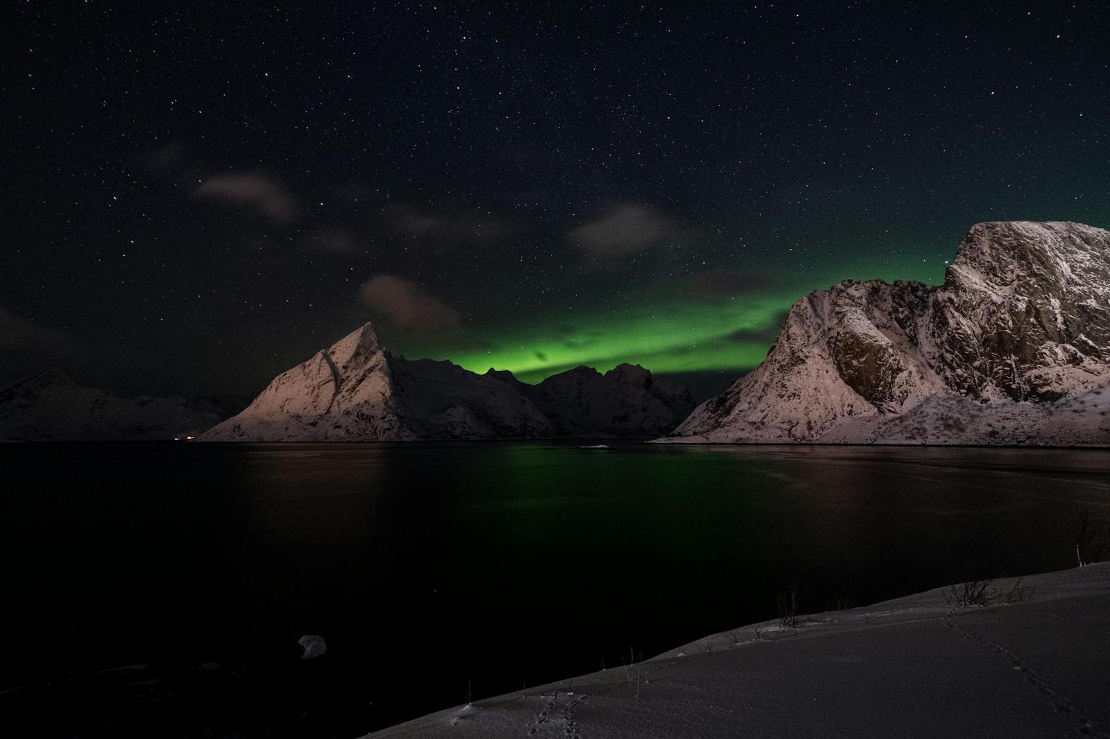 The northern lights glowing green over snow-covered coastal peaks at night