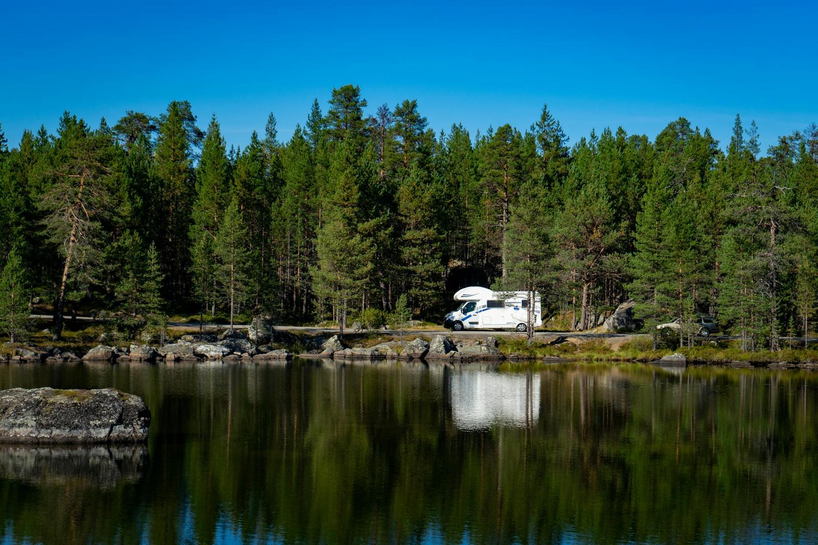 A camper van parked among pine trees beside a still lake