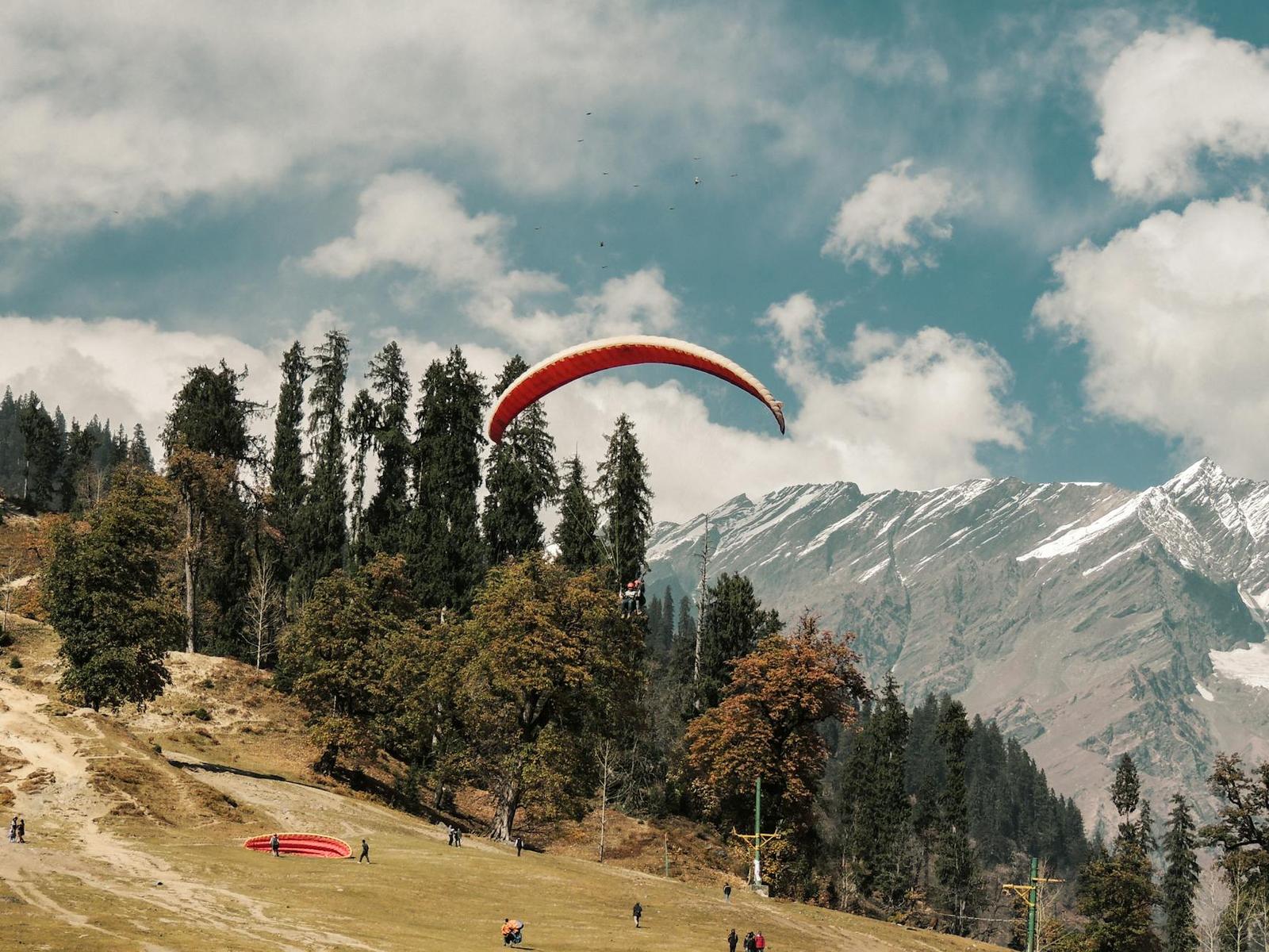 A paraglider descending over a grassy slope with snow peaks beyond