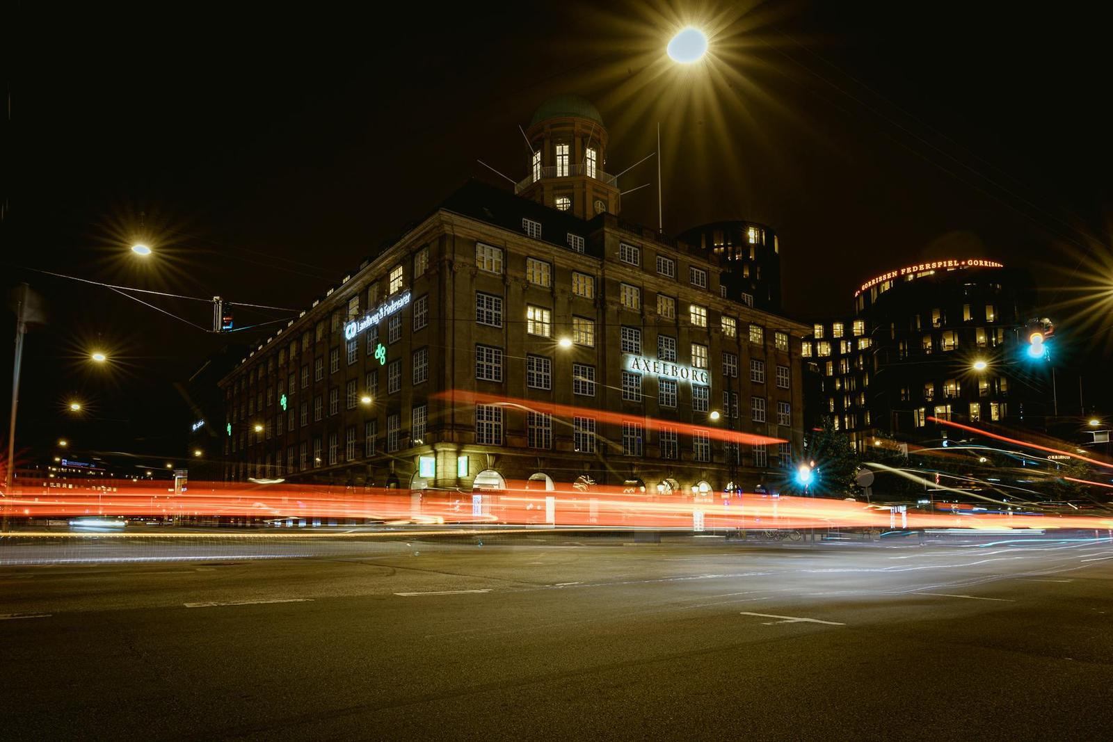 Light trails streak past a lit brick building on a city street at night