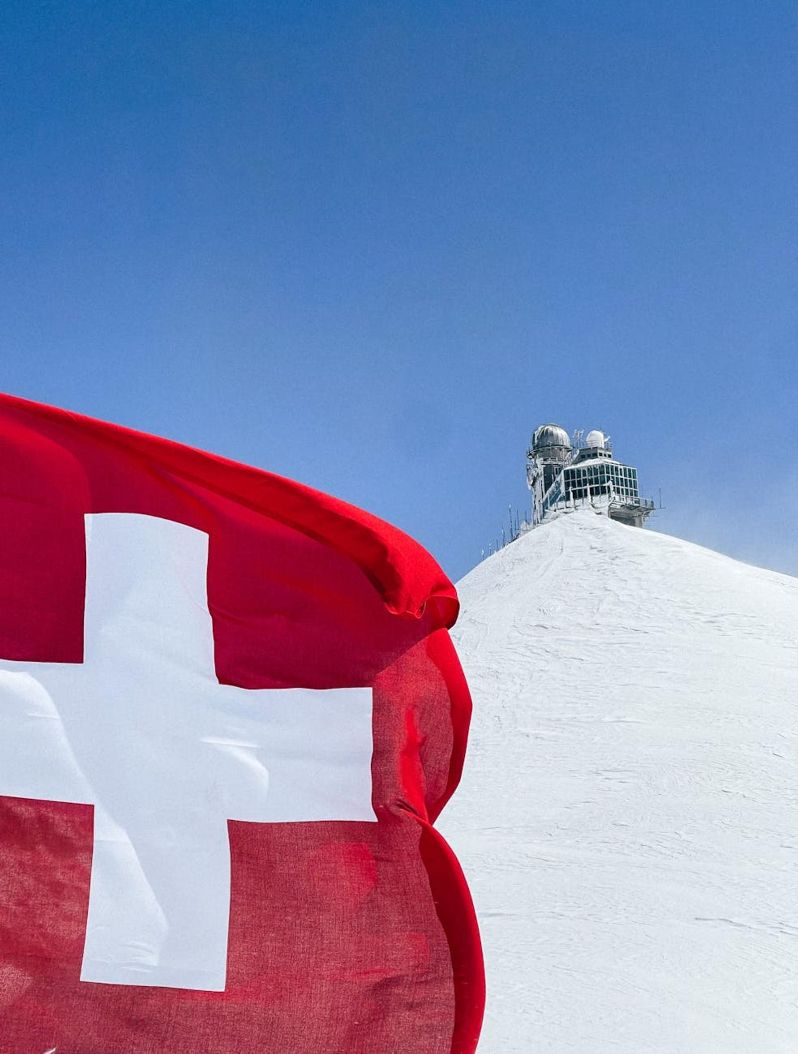 Swiss flag waving in front of the Sphinx Observatory on a snowy peak, under a clear sky