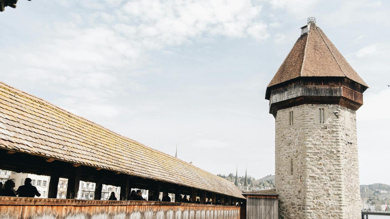 A stone tower with a pointed roof beside a long wooden covered bridge