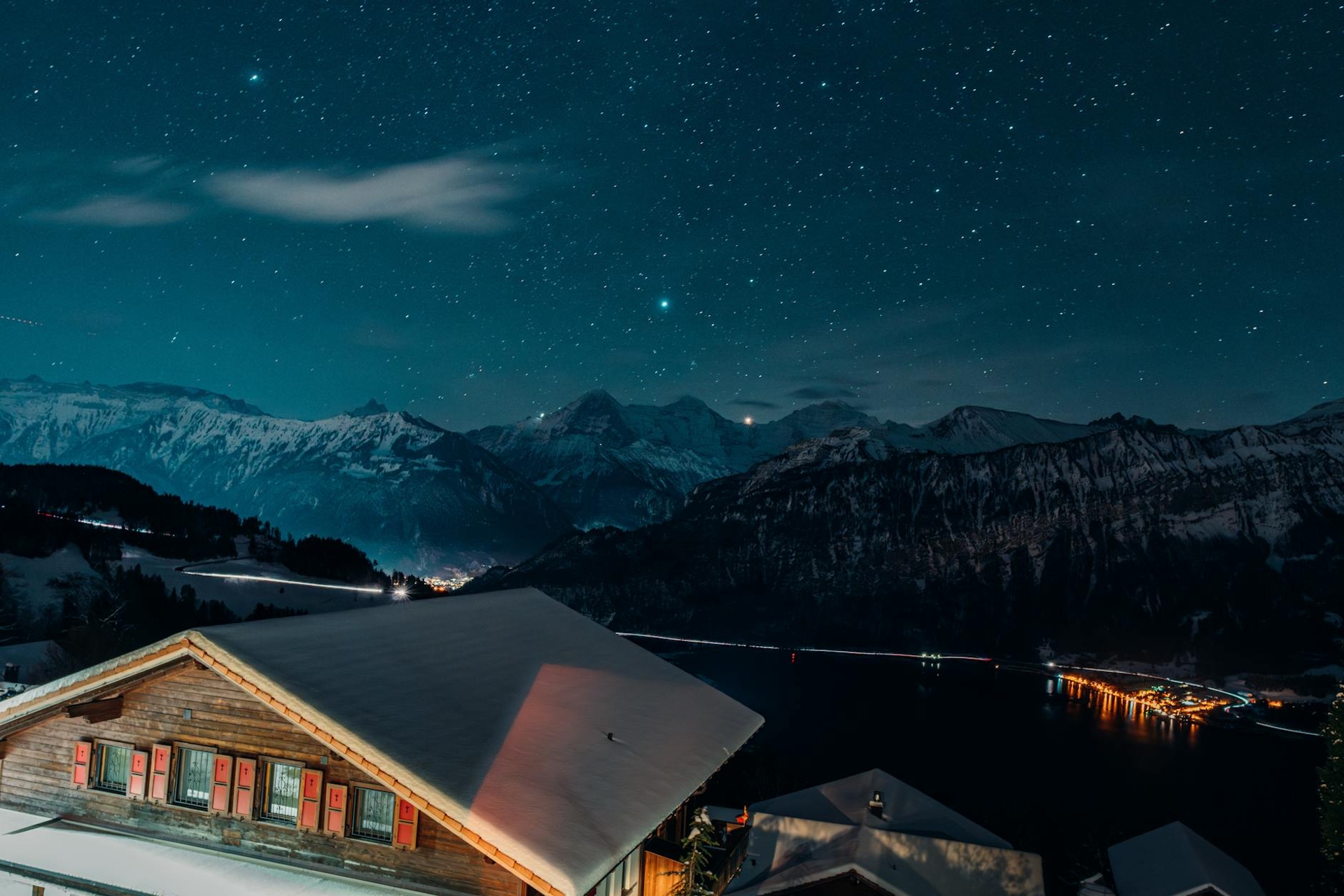 A starry night sky over snow-dusted mountains and a lakeside village with lit windows