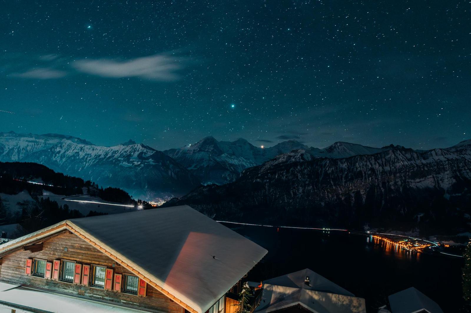 A starry night sky over snow-dusted mountains and a lakeside village with lit windows