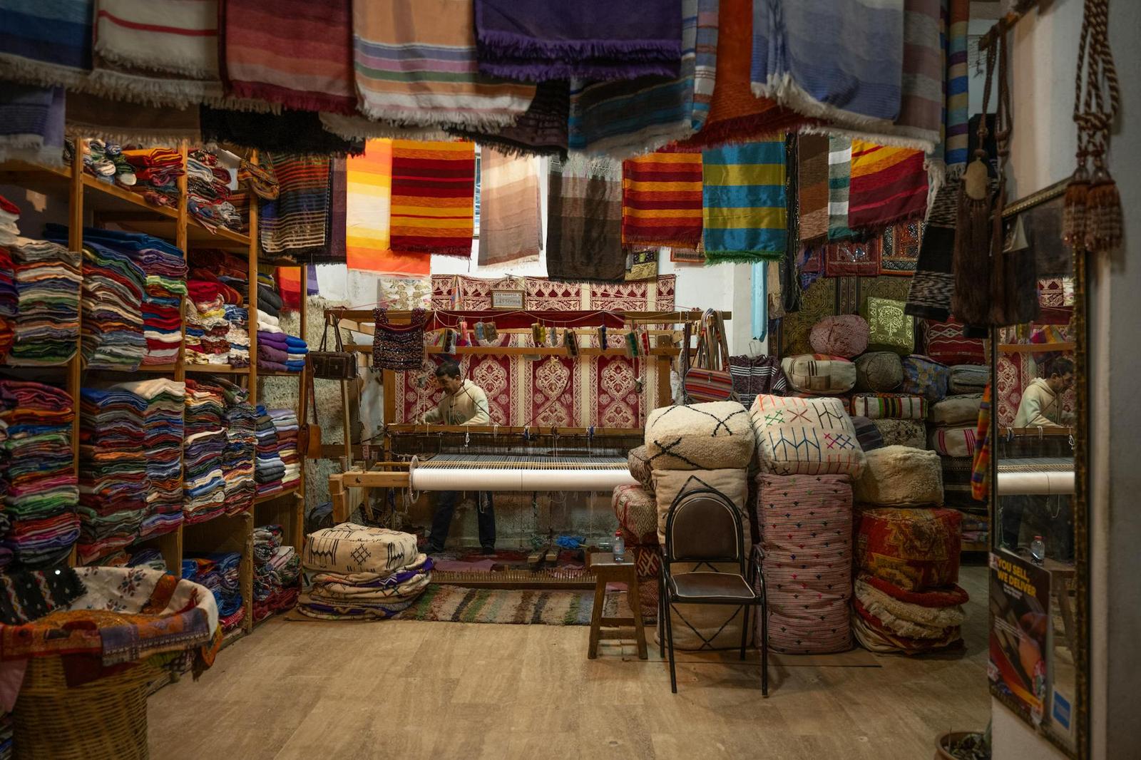 A textile shop stacked with folded rugs and blankets around a weaver at a loom