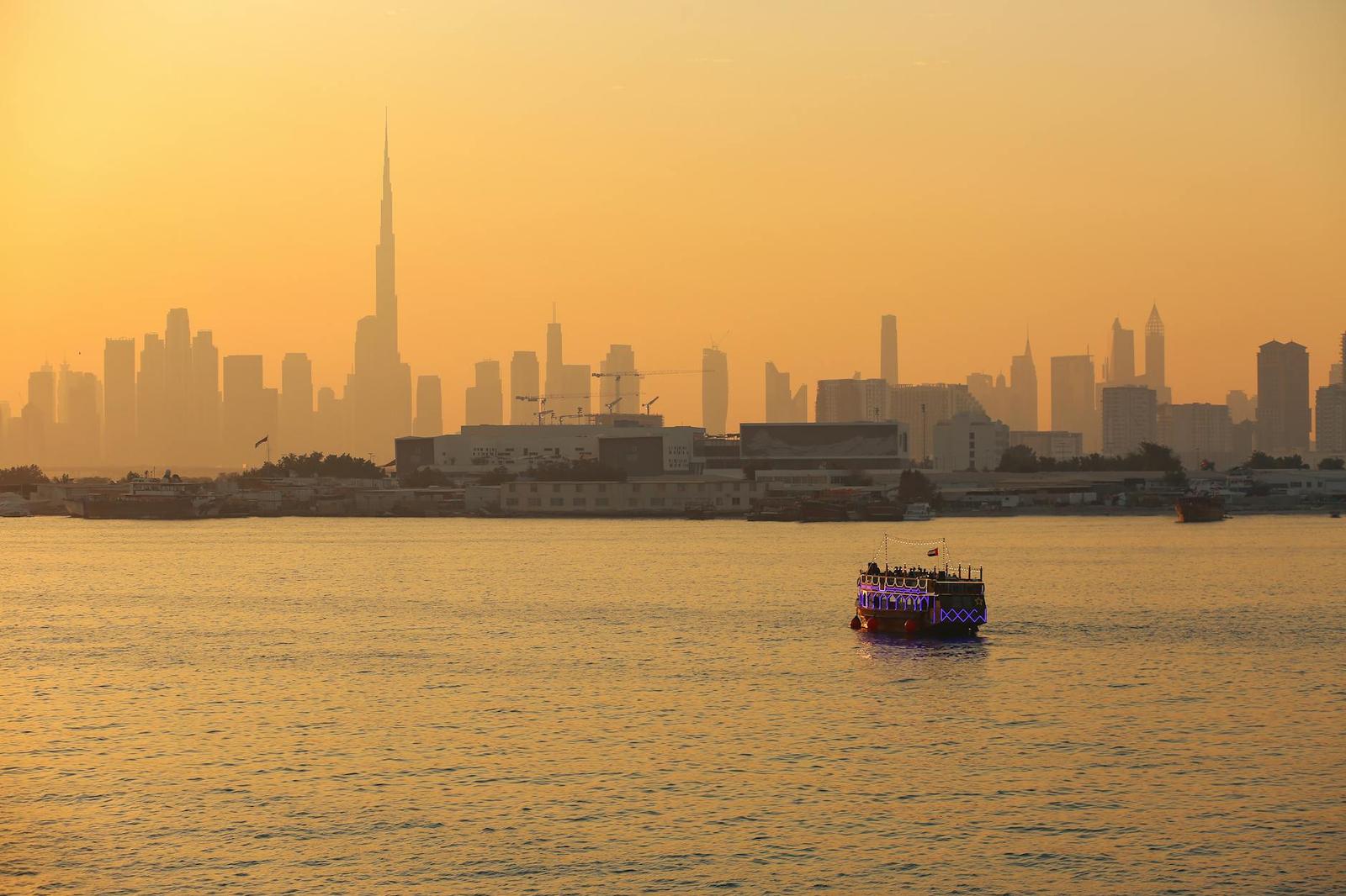 A lit traditional cruise boat on a river at sunset with a spired skyline behind