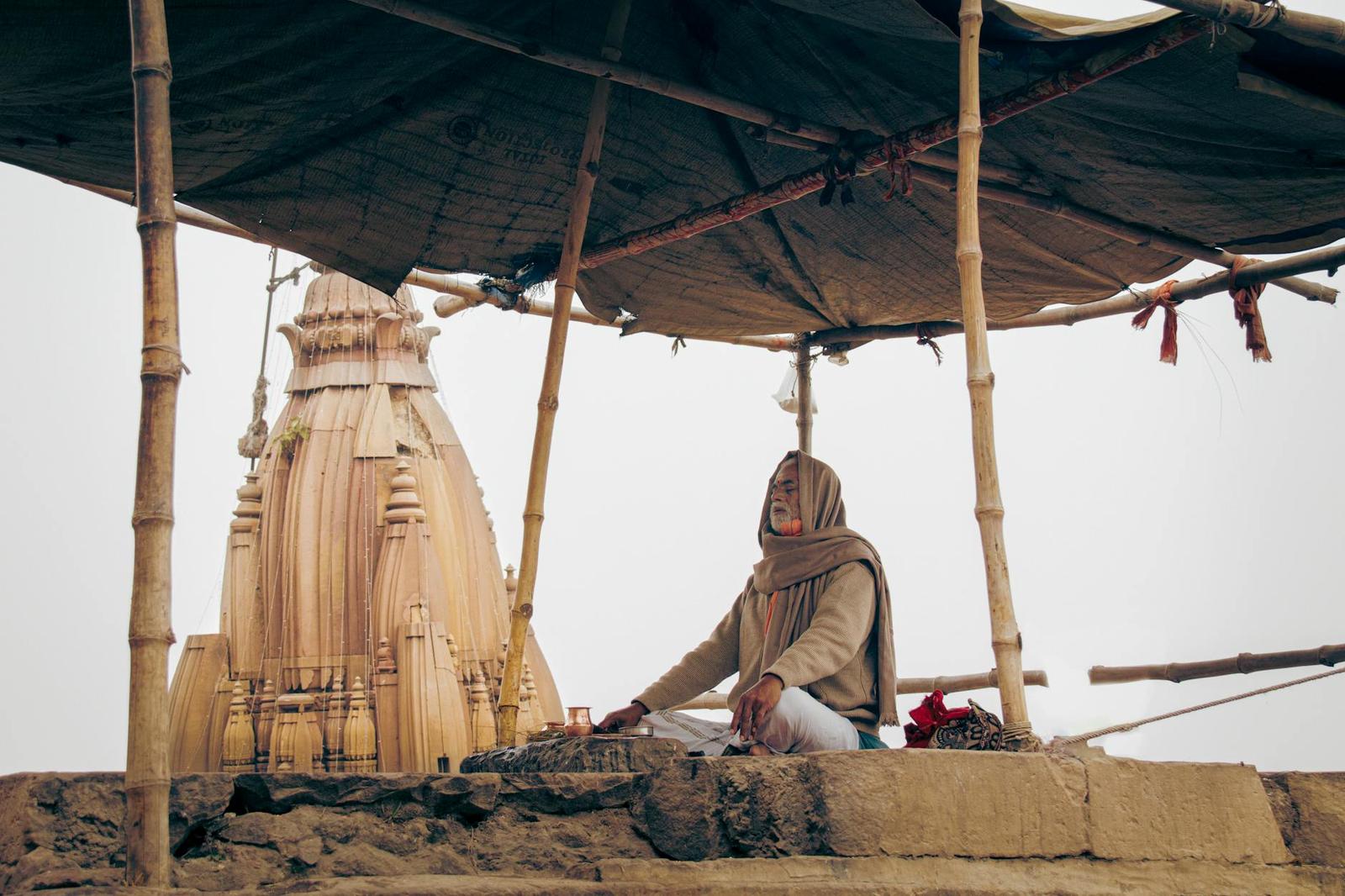 A person sitting cross-legged under a tarp canopy beside a small carved temple model