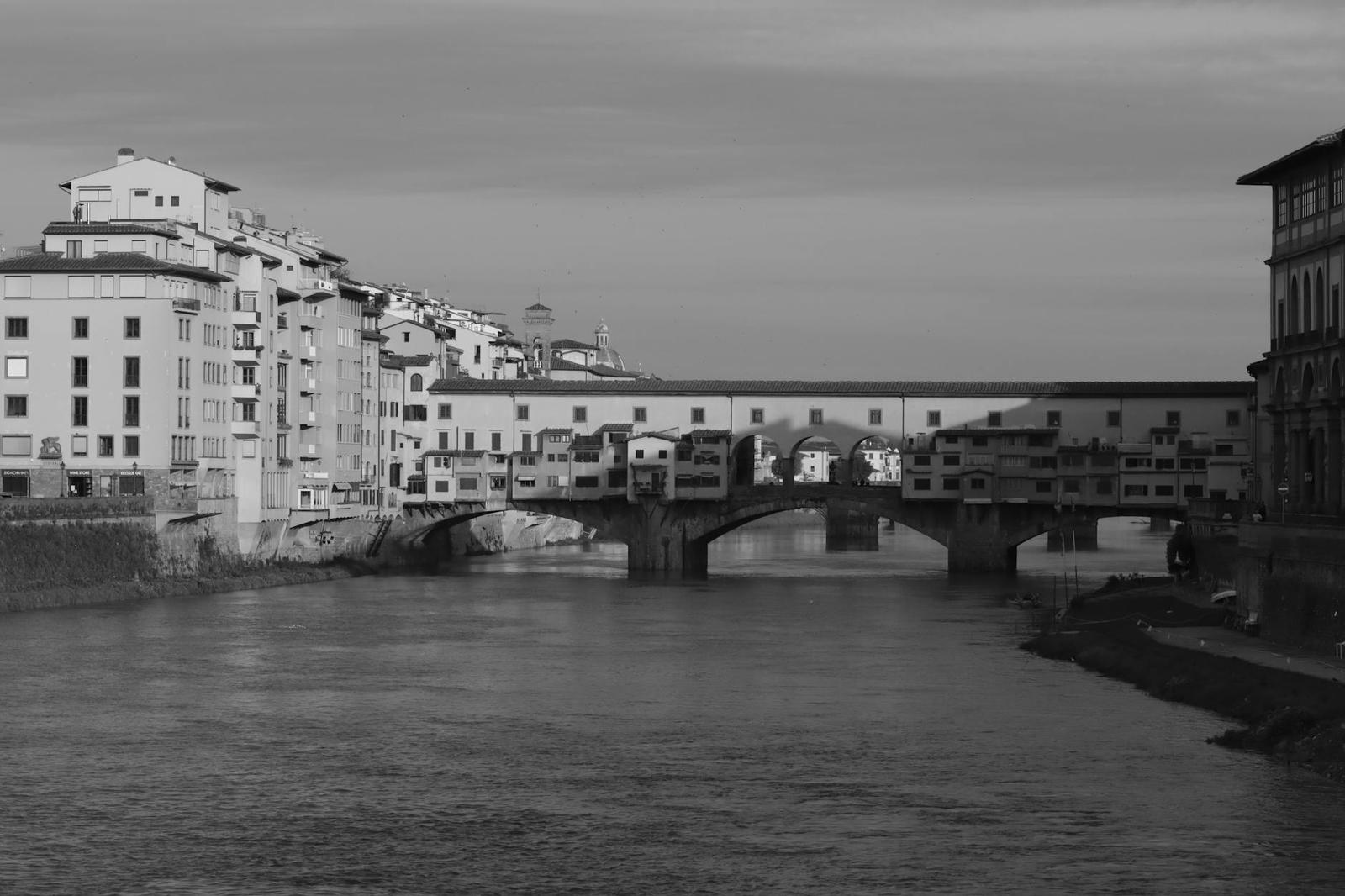 Black and white view of a covered stone bridge spanning a river at dusk