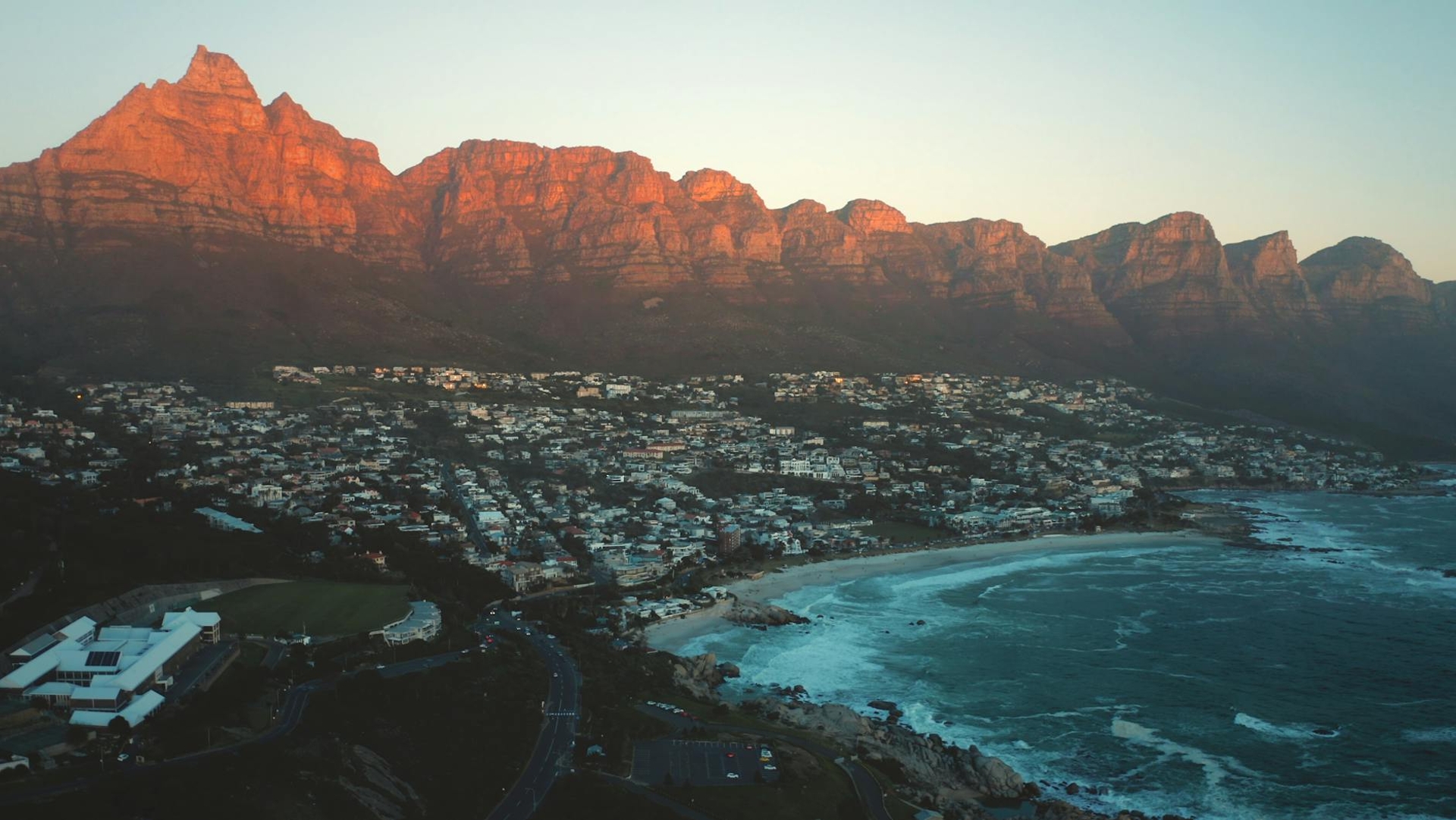 Jagged mountains glow red at sunset above a coastal town and beach