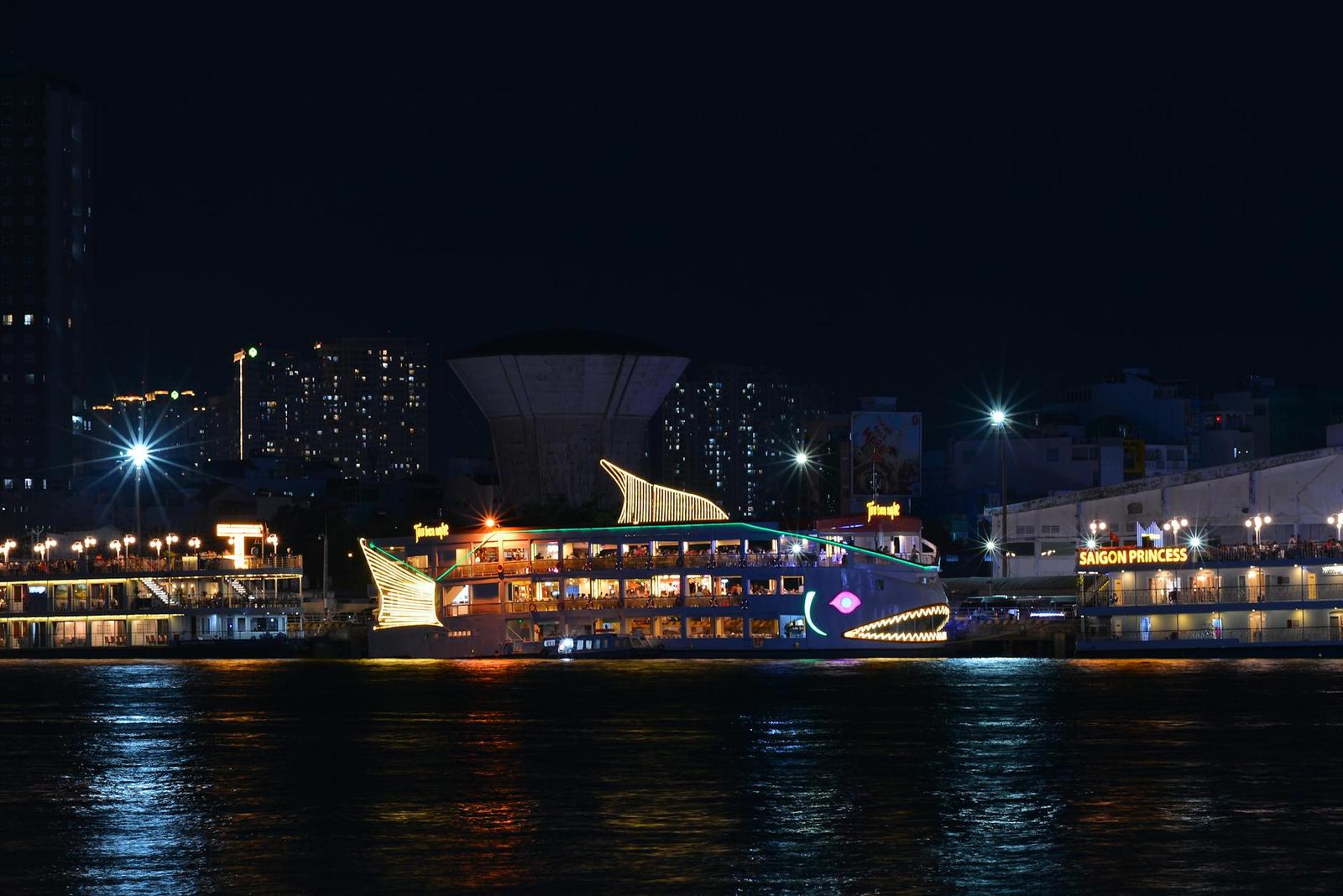 A dragon-shaped illuminated boat cruising a river past a lit-up city skyline at night