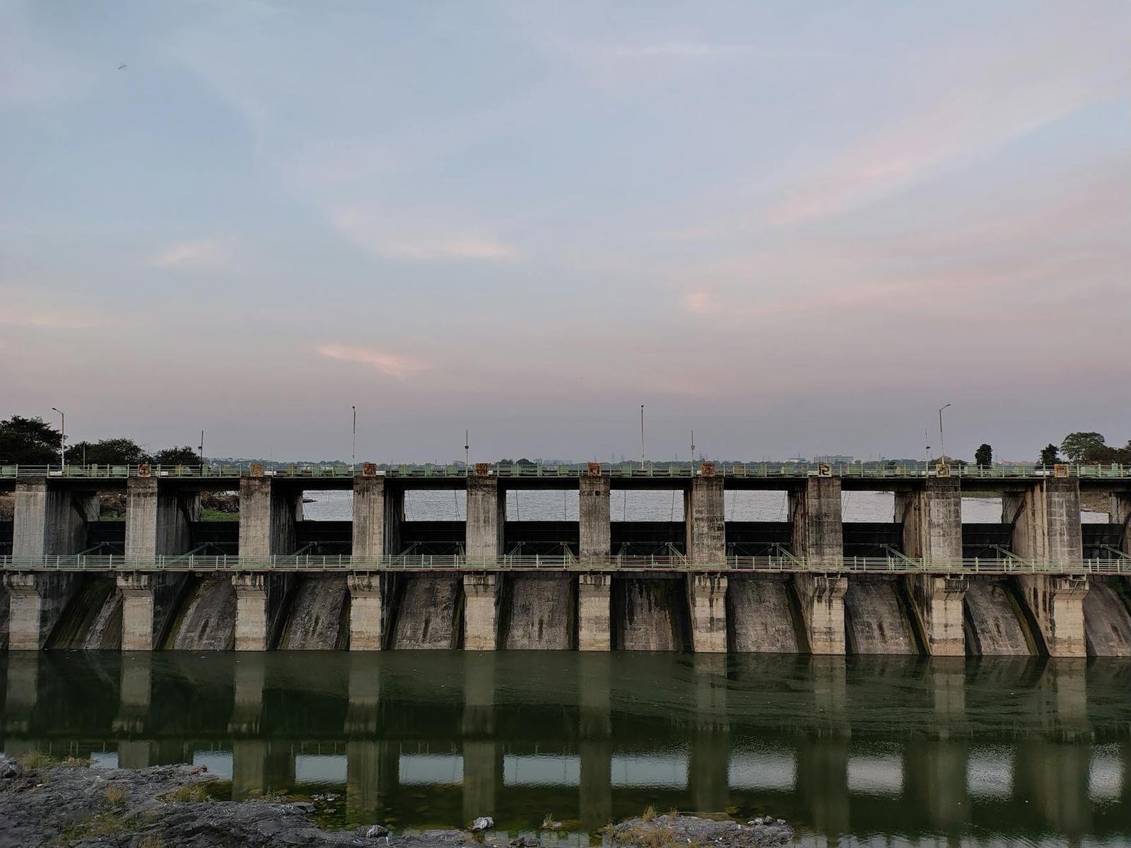 A long concrete river dam with multiple gates reflected in still water at dusk