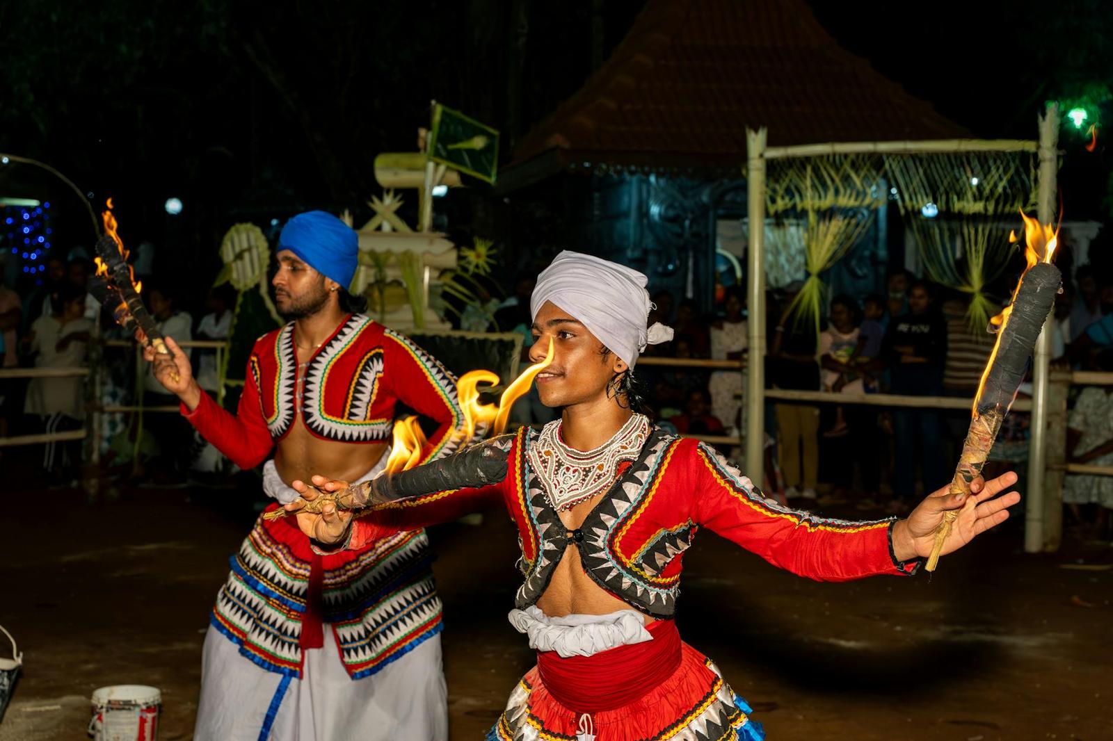 Two dancers in ornate red costume perform with flaming torches at night