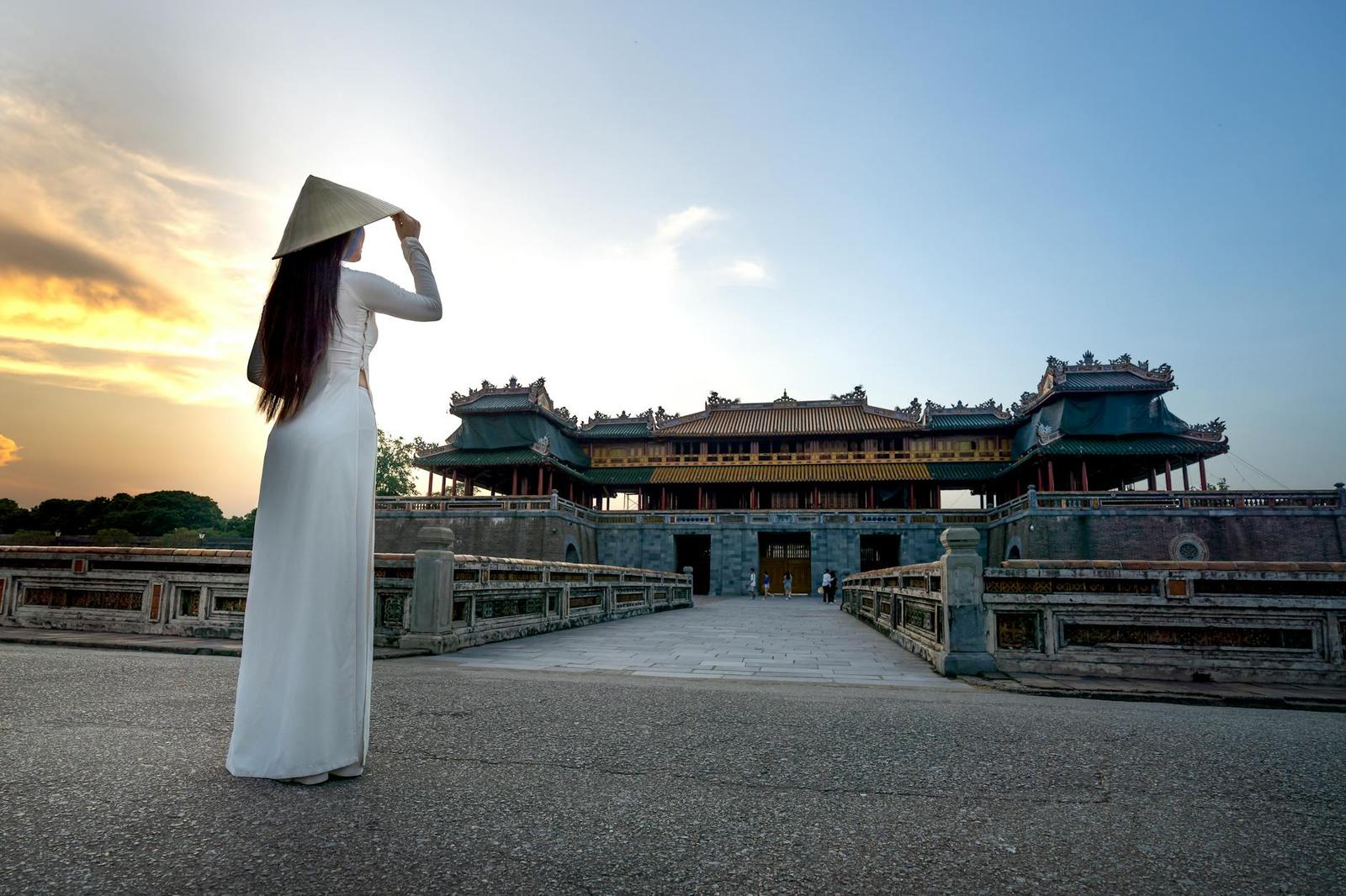 Woman in a white ao dai facing an ornate citadel gate at sunset