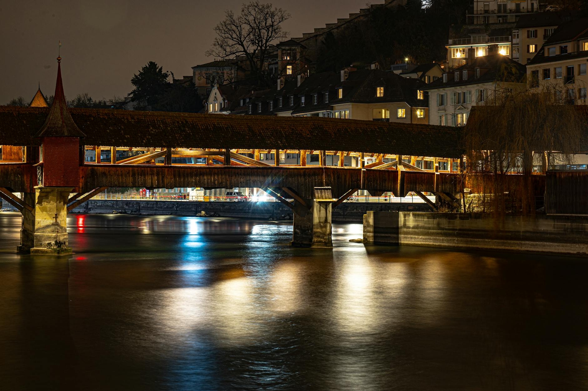 A covered wooden bridge lit at night reflecting on a calm river