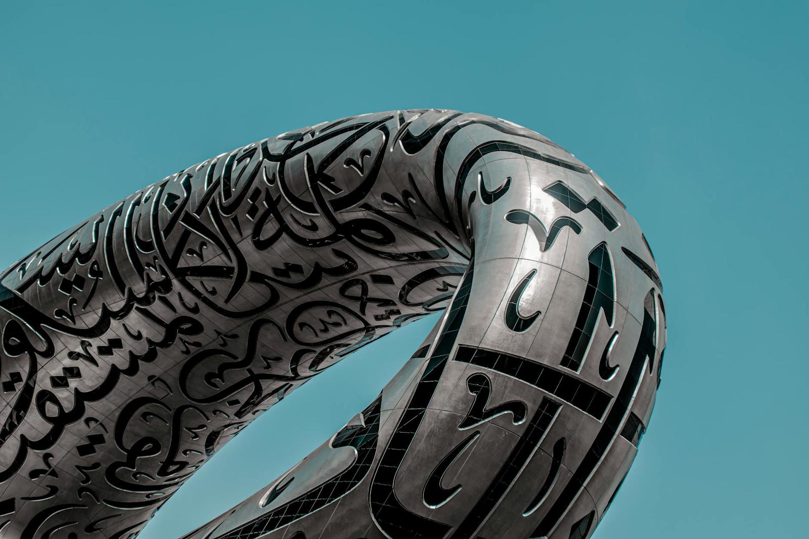 A close-up of a curved metal structure covered in flowing calligraphy script against a blue sky