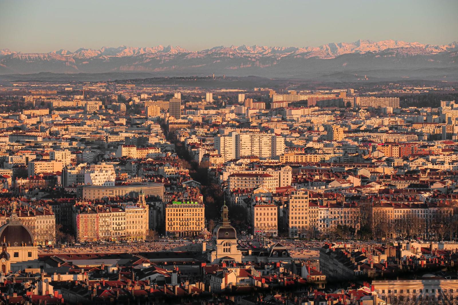 A dense city skyline at sunset with distant snow-capped peaks on the horizon