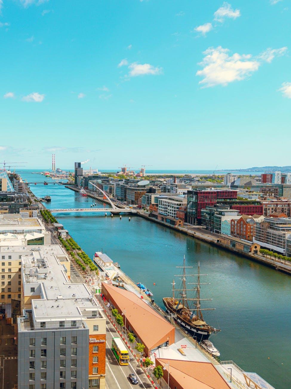 Aerial view of a riverside city with a tall ship moored beside red-roofed warehouses