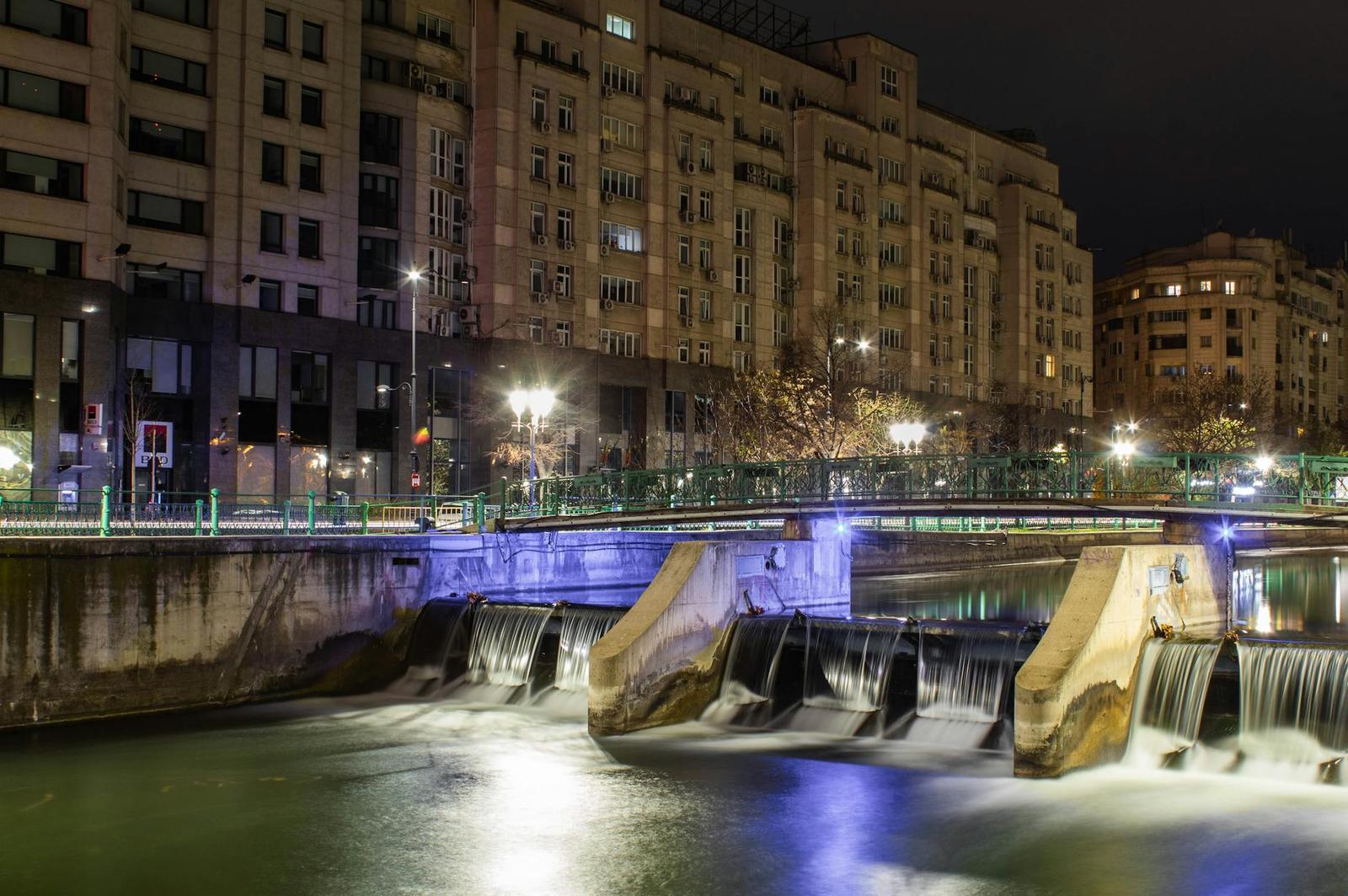 A lit stepped weir on a city river at night flanked by apartment blocks